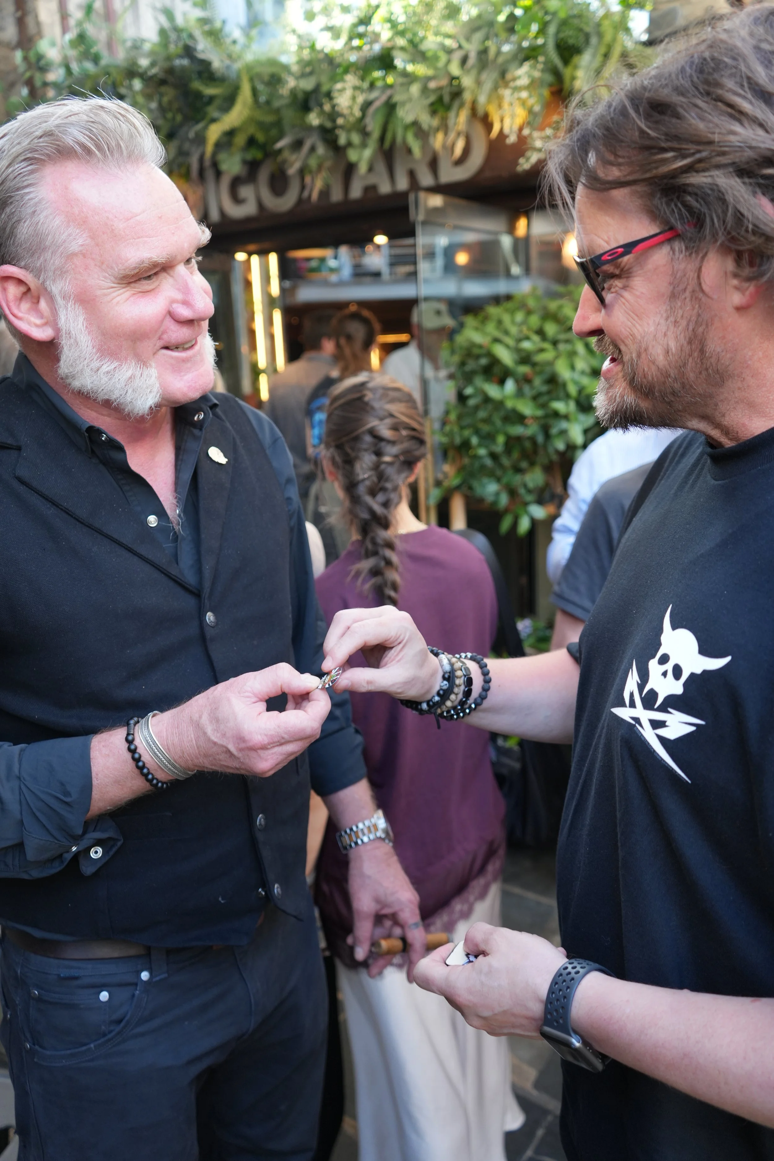 Two men are smiling and engaging in a conversation outdoors, with several people in the background near a building with a sign that reads 'GOTARD'. They are exchanging or holding jewelry or small items, and one man is wearing a black shirt with a sku