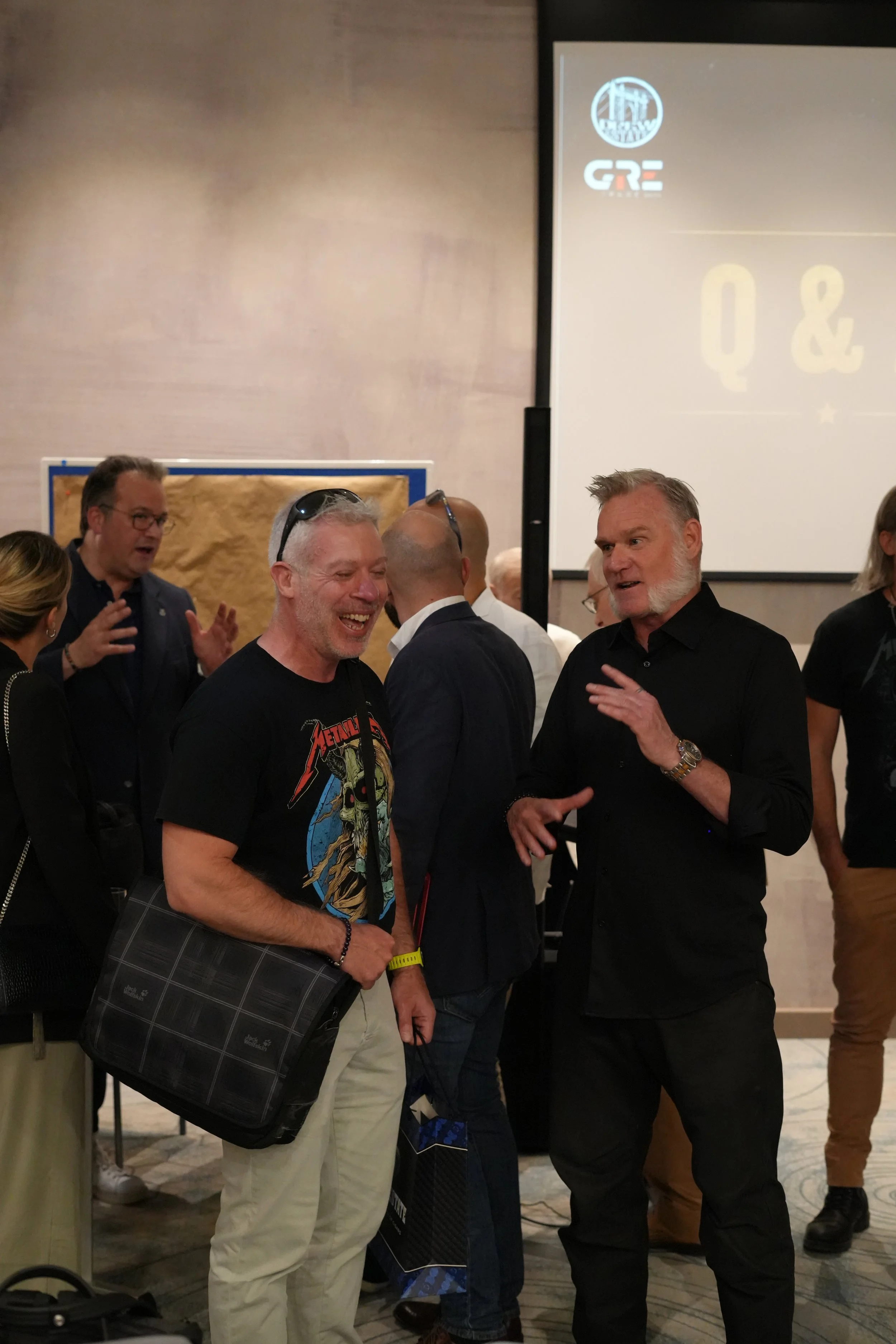 Group of people at an indoor event, engaged in conversation, with a large screen displaying logos in the background.
