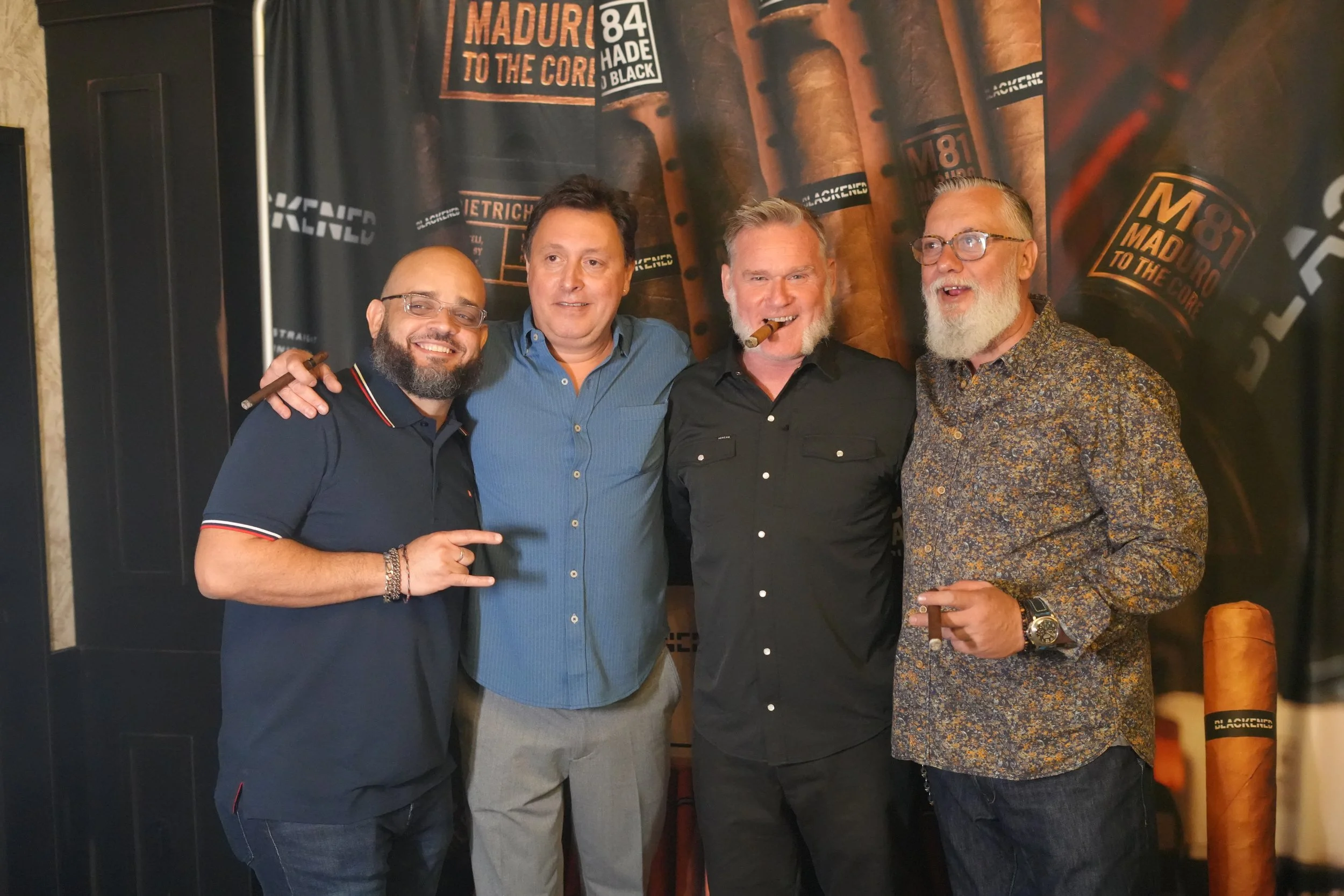 Four men stand together in front of a backdrop with cigar and tobacco branding, smiling. The man on the far left is wearing glasses and a navy polo shirt, the second man is wearing a blue button-up shirt, the third man has a cigar in his mouth and is
