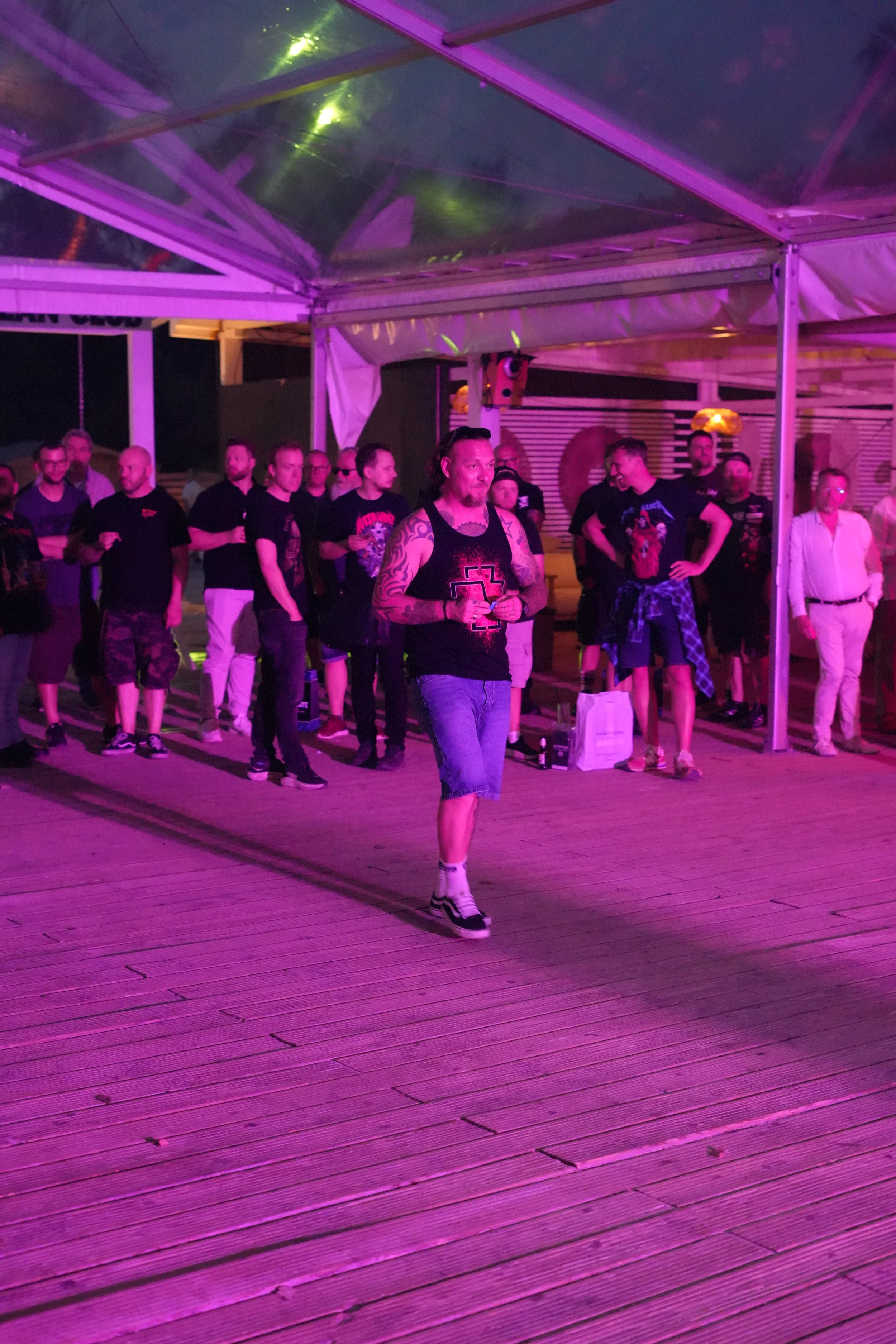 A man with tattoos on his arms wearing a black tank top, shorts, and sneakers, walking on a wooden dance floor under pink and purple lighting. He is holding a drink and appears to be at a social event with a group of people in the background.