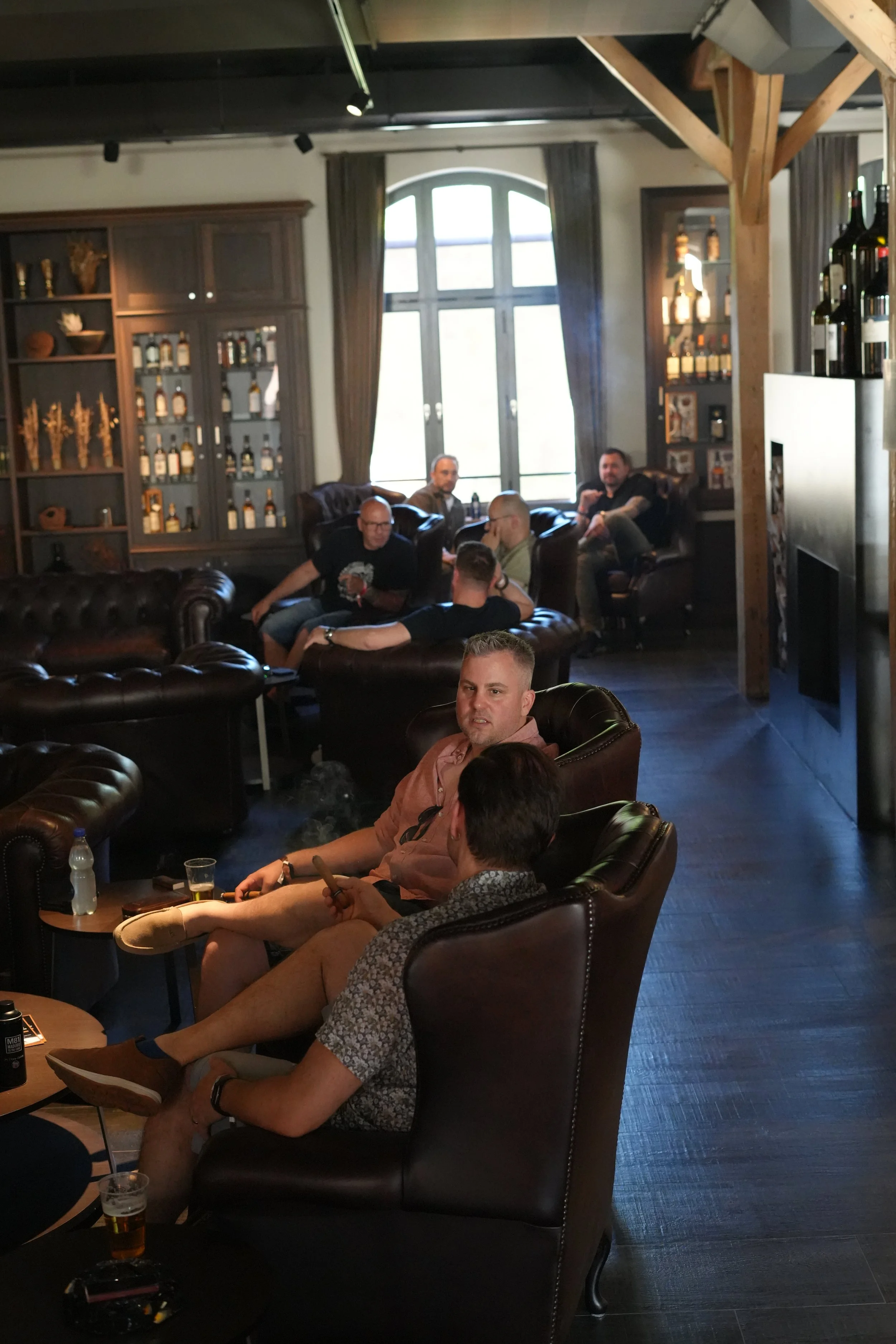A group of men seated in a cozy bar or lounge area with dark leather chairs and wooden shelves filled with bottles and decor. Large windows with curtains are visible in the background, and some men are engaged in conversation while others are using t