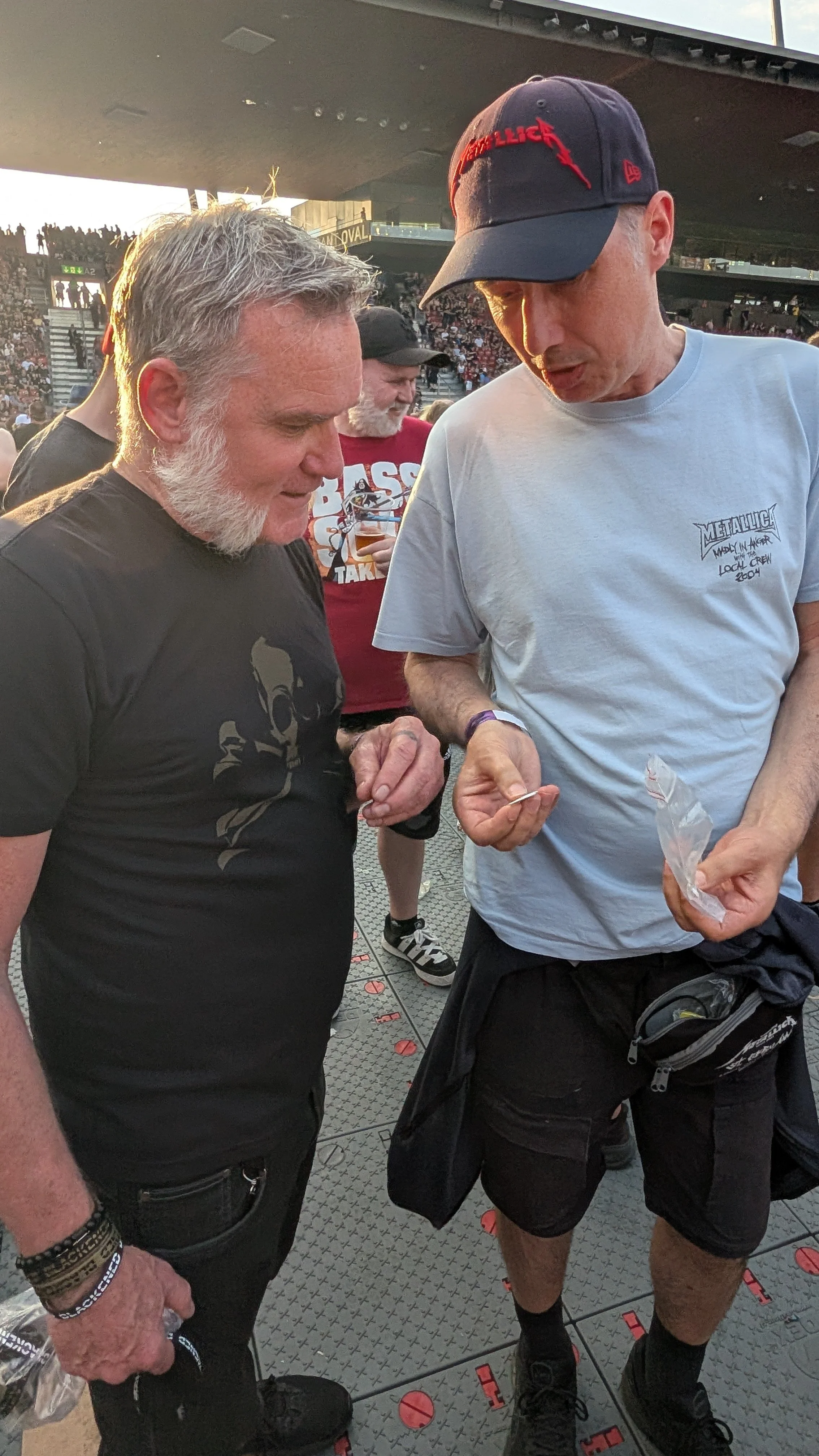Two men are looking at something in each other's hands at a Metallica concert. One man has gray hair and a beard, wearing a black shirt, and the other man is wearing a white t-shirt, a cap with red logo, and black shorts.