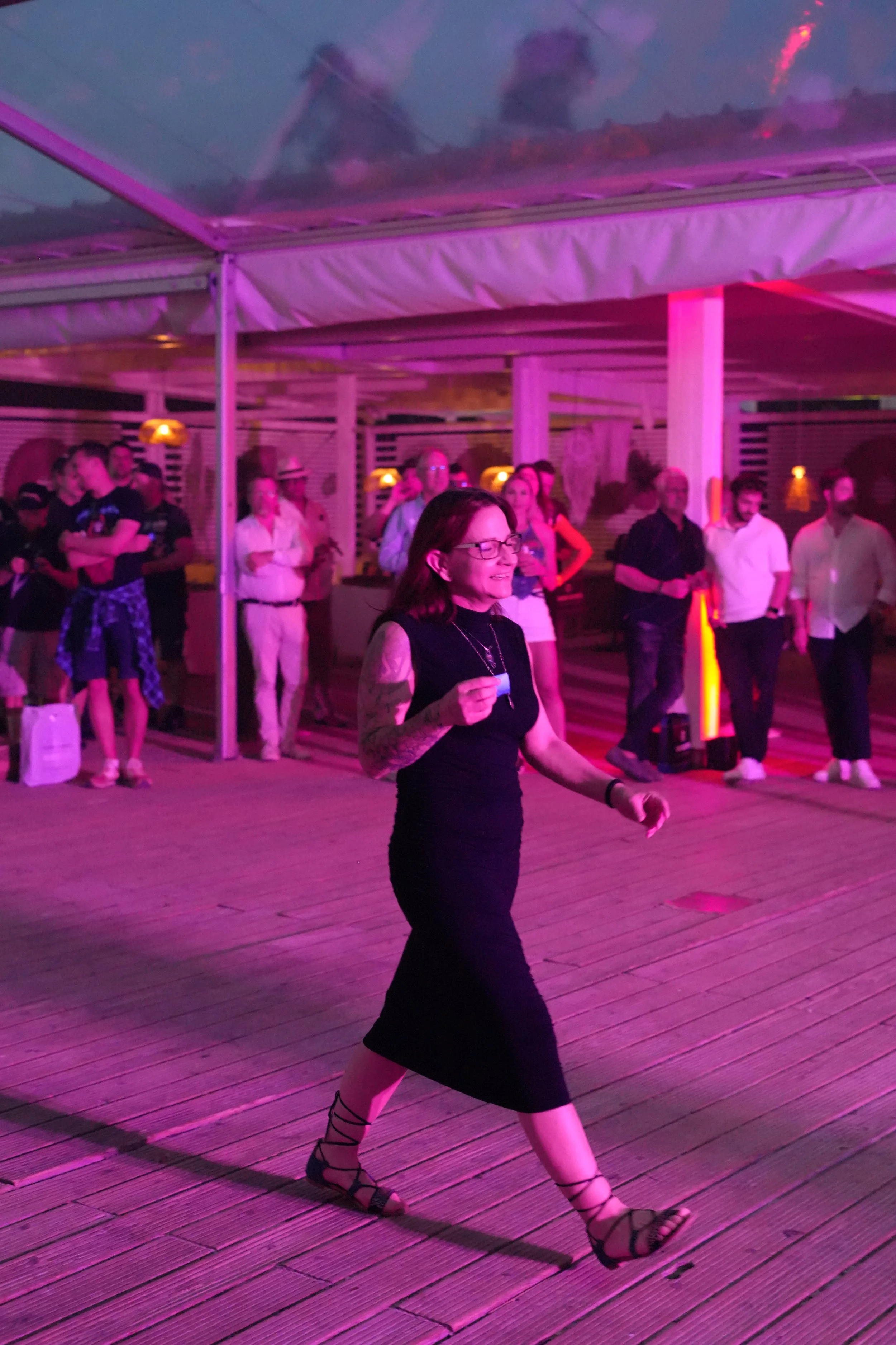 A woman with glasses and tattoos on her arms dancing on a wooden deck under purple lighting. Several people are in the background, gathered and watching, in an outdoor or semi-outdoor setting with a modern, wooden structure.