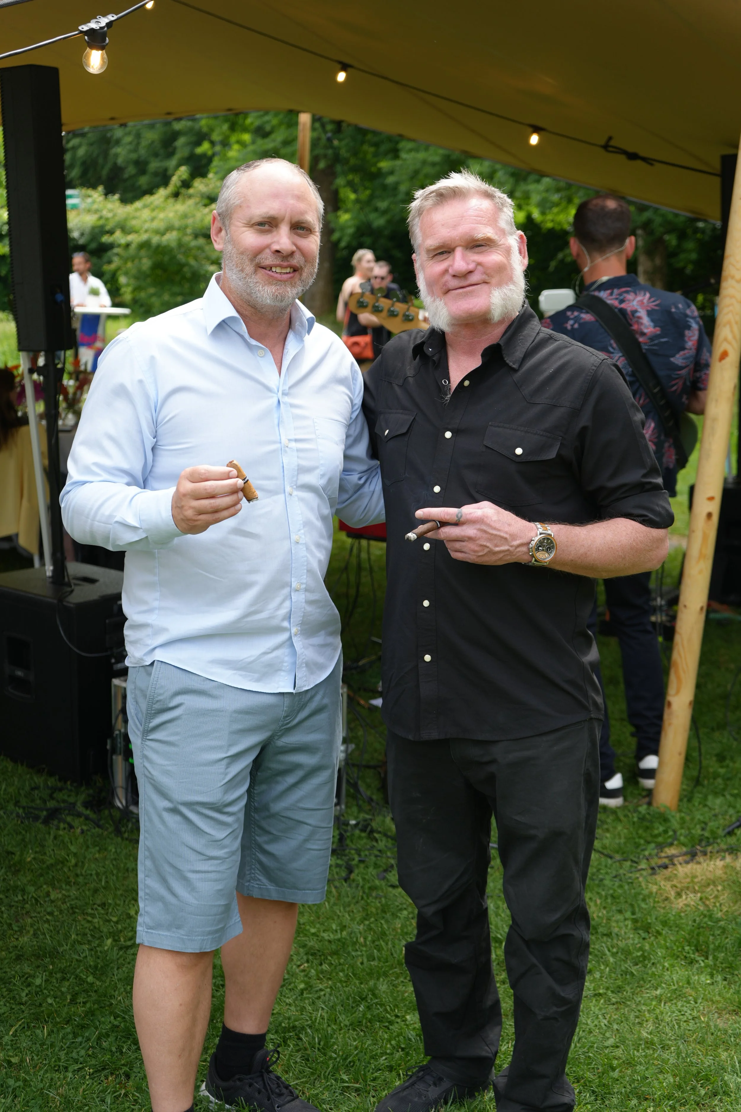 Two men standing outdoors under a yellow canopy, smiling at the camera, with one holding a small cigar or cigarette, and the other holding a cigarette. The background shows other people and green trees, indicating an outdoor event.