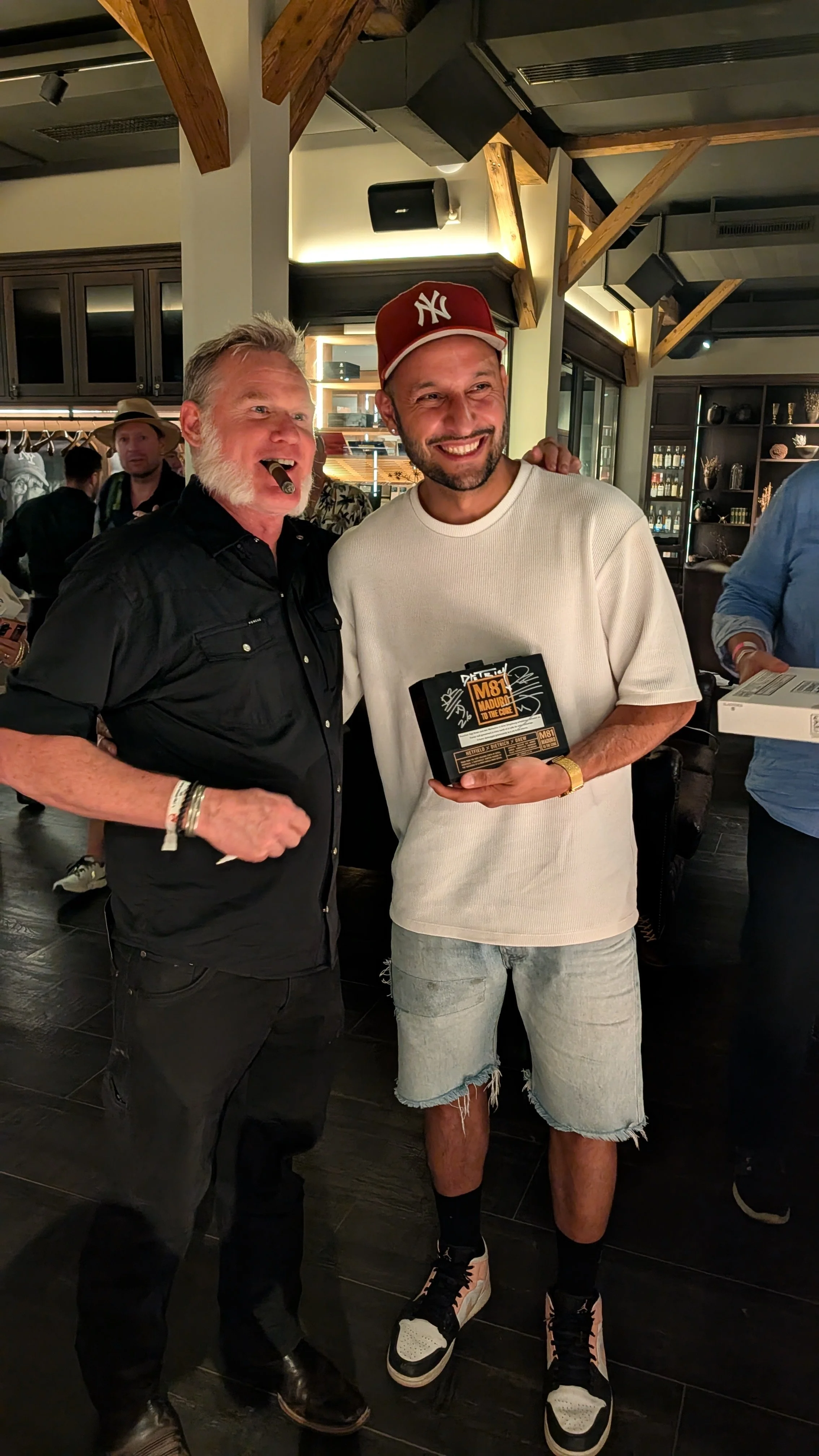 Two men smiling, one holding a package. One man has gray hair and beard, wearing black shirt and accessories. The other man has a beard, wearing a New York Yankees cap, white t-shirt, ripped denim shorts, and sneakers, with a gold watch.
