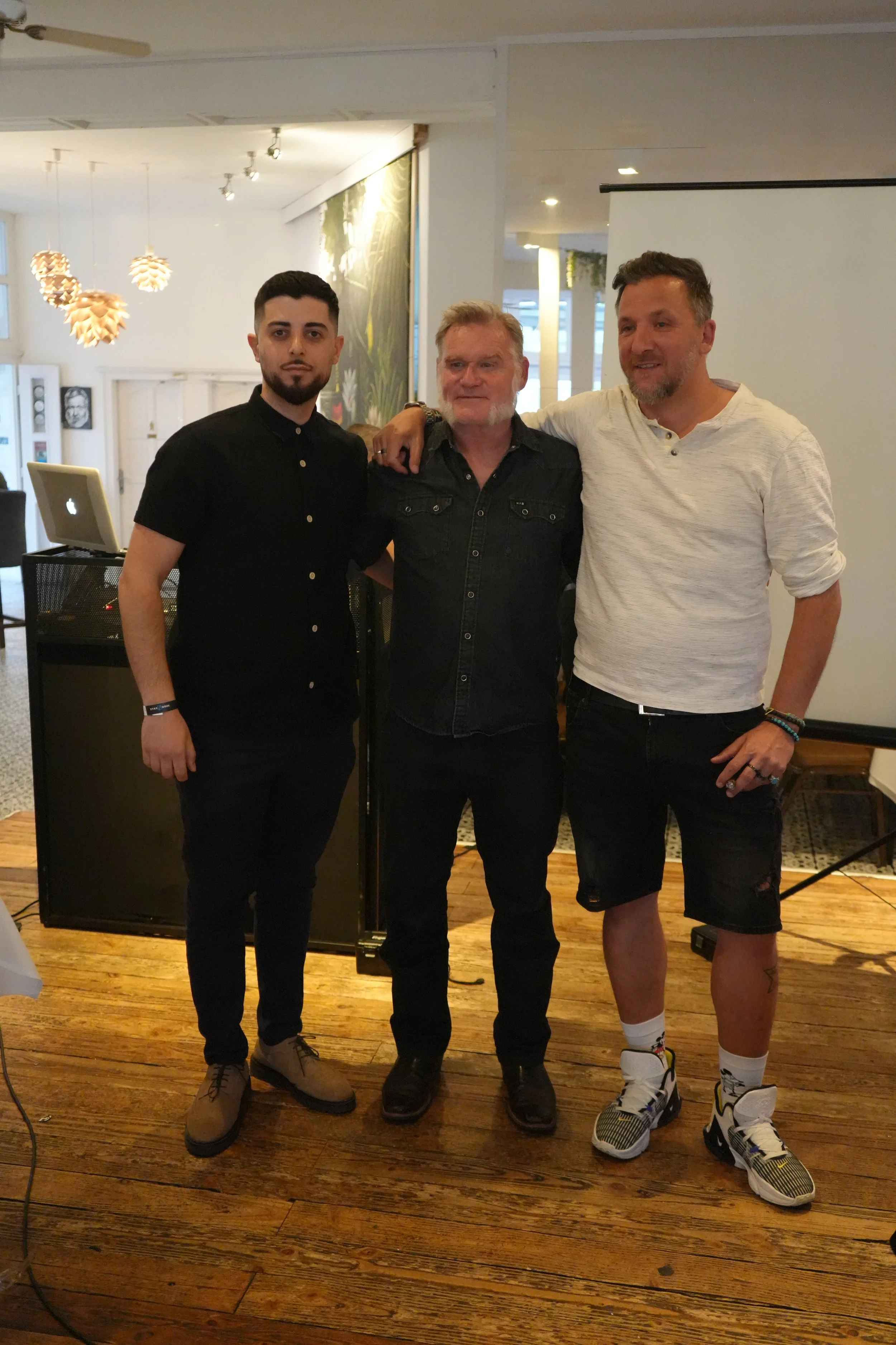 Three men standing together inside a building with a wooden floor. They are posing for the photo, with the man in the middle having his arms around the other two. The man on the left is young, with dark hair and a beard, wearing a black shirt and dar