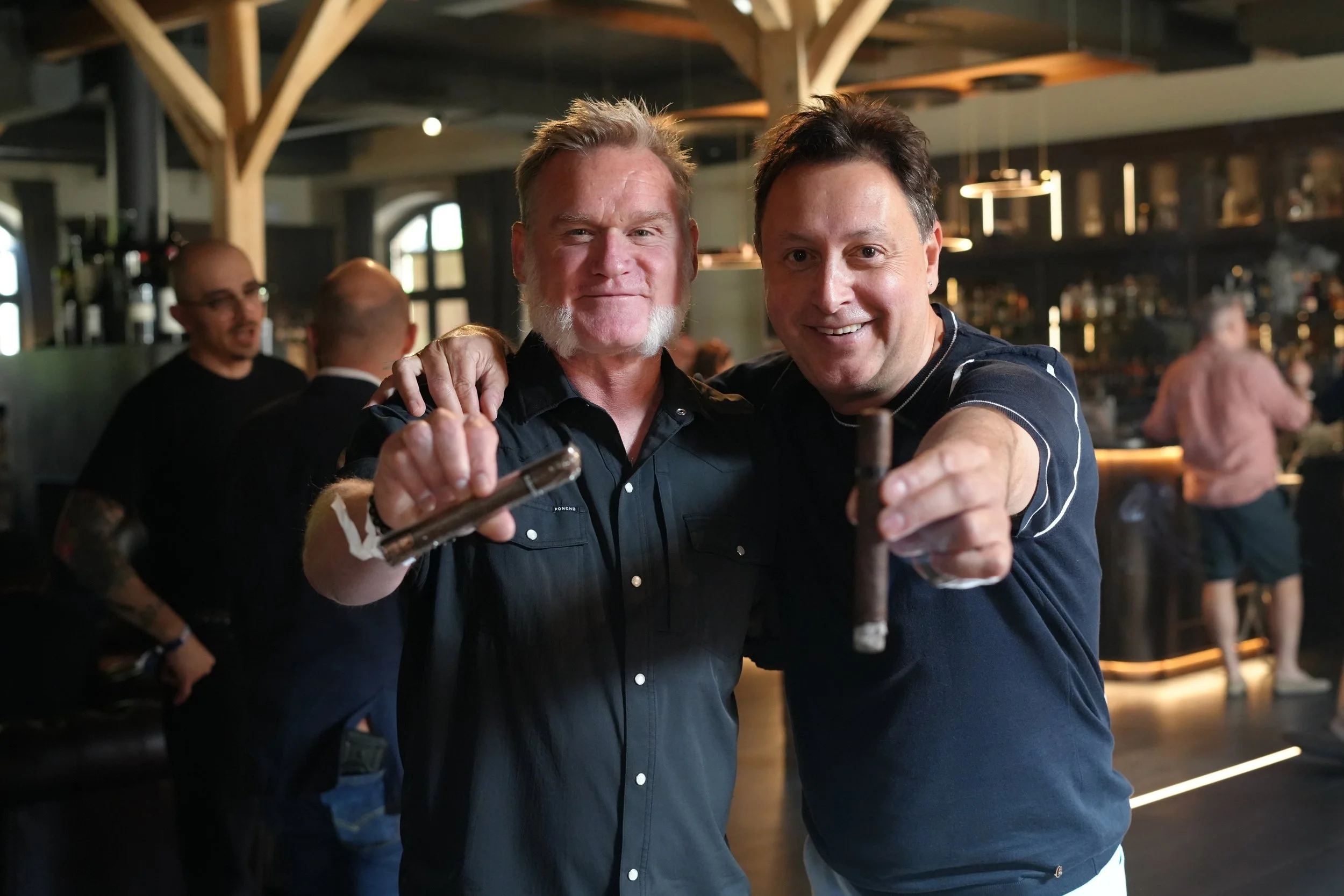 Two men smiling and posing for a photo in a lively indoor setting, possibly a bar or restaurant, with other people and dim lighting in the background.