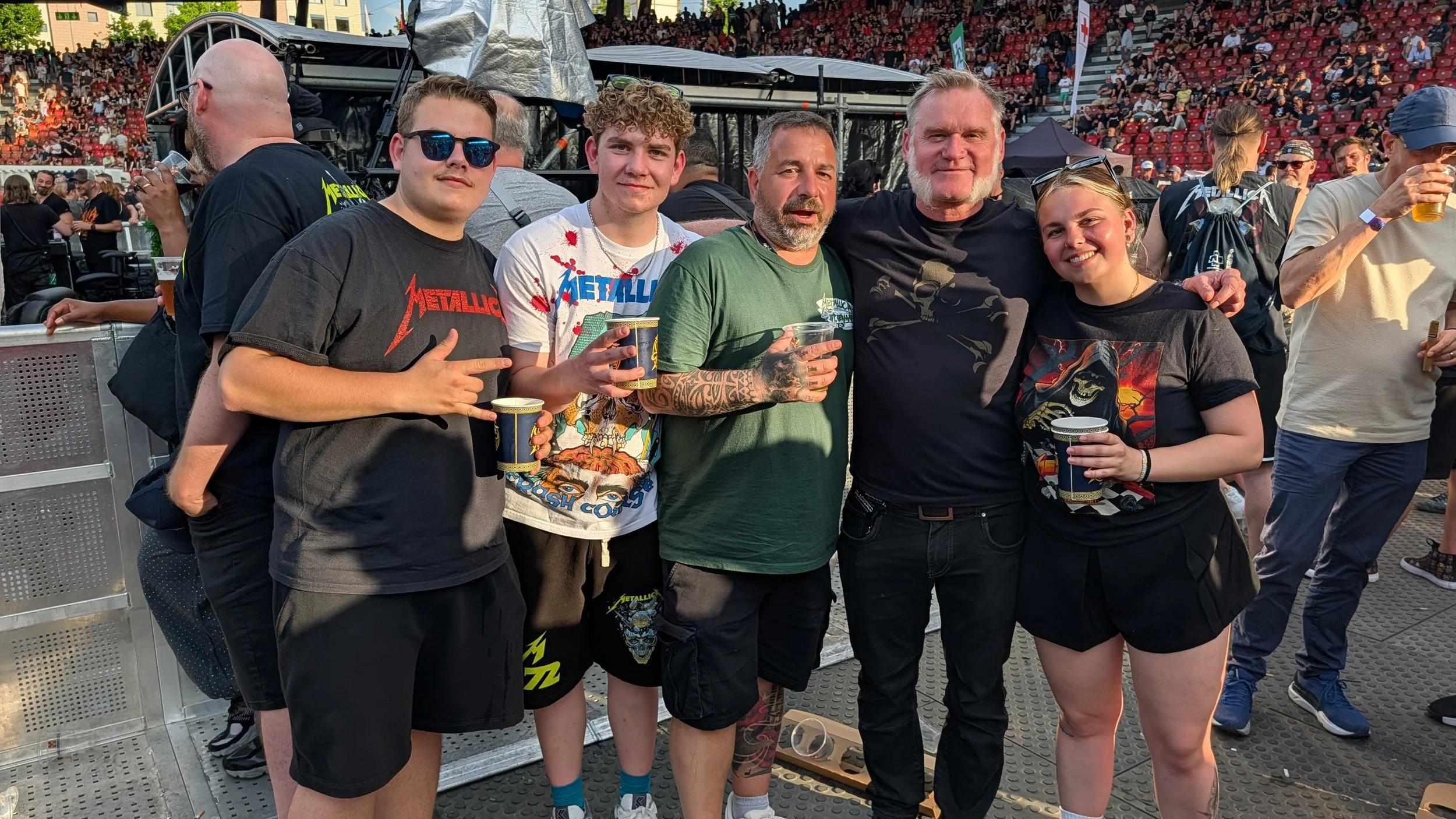 Group of five people, four men and one woman, standing together at a Metallica concert with a crowd in the background. They are holding drinks and smiling, with some wearing Metallica band shirts and sunglasses.