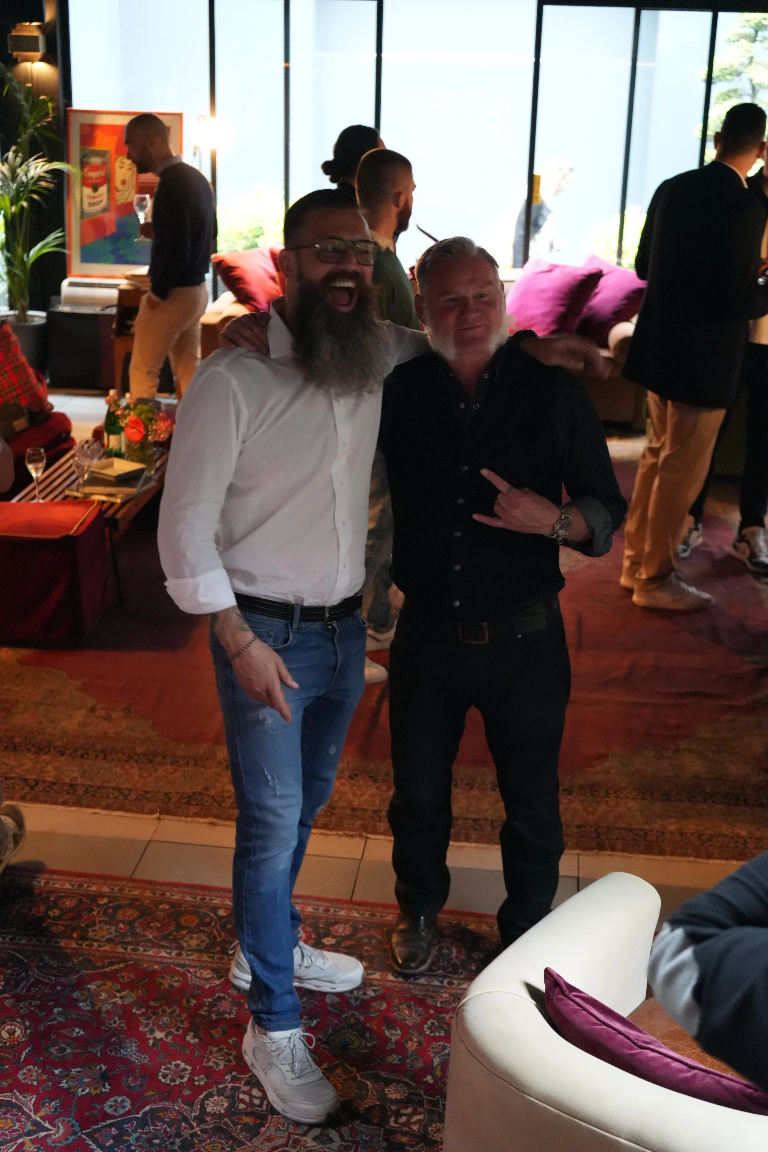 Two men standing inside a modern, stylish room with large windows. One man wears a white shirt and jeans, smiling with his mouth open, and the other wears a black shirt with an arm around the first man. The room is decorated with colorful pillows, ar