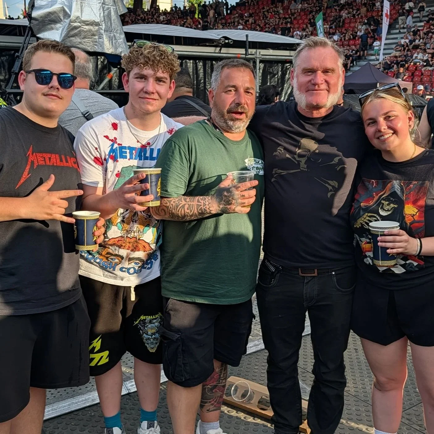 Group of five people at a Metallica concert holding cups, with an audience seated in the background.