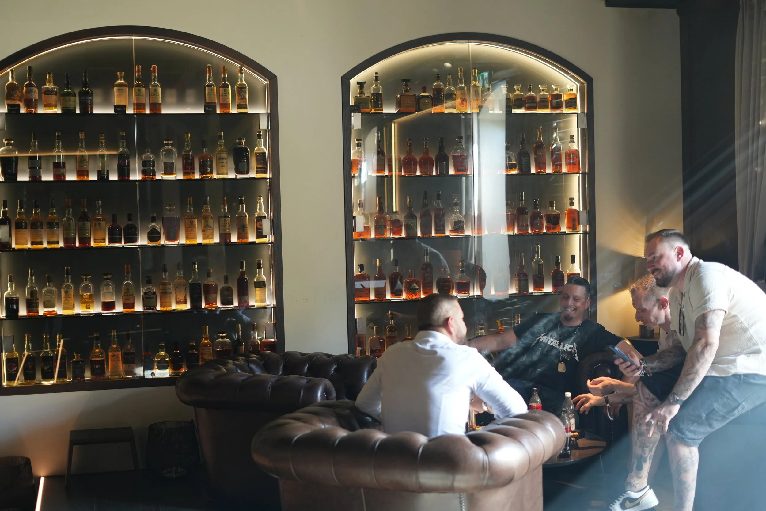 A group of people sitting and standing in a room with two large shelves displaying bottles of alcohol on illuminated shelves.