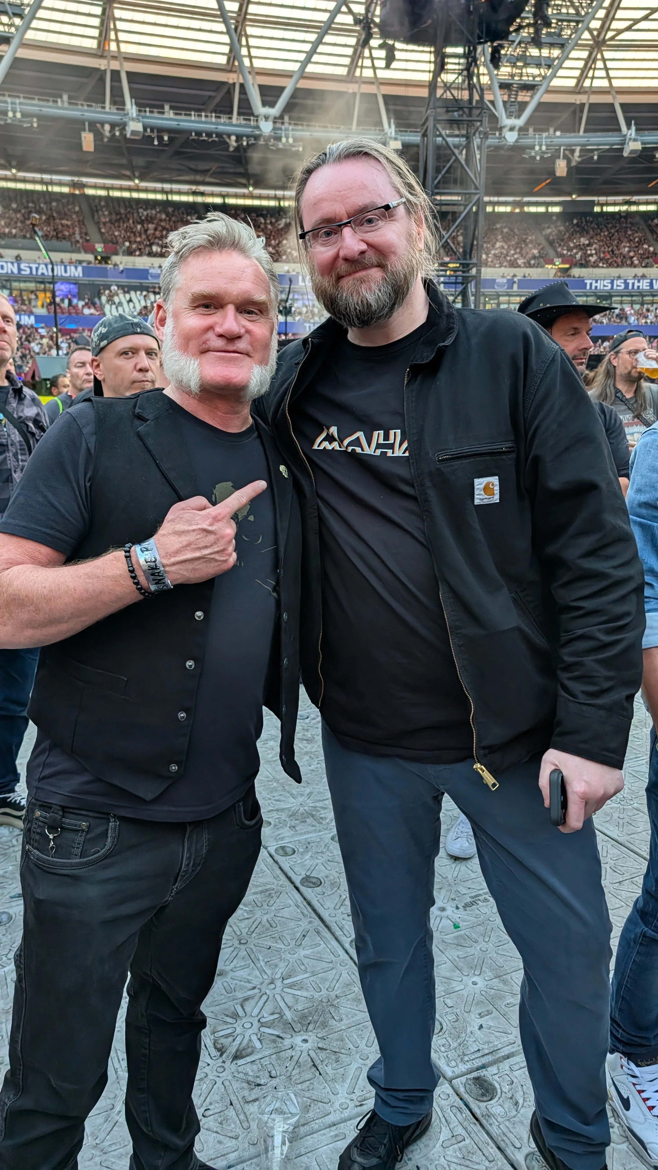 Two men standing together at a concert, one pointing at the other's chest, in a stadium filled with people.