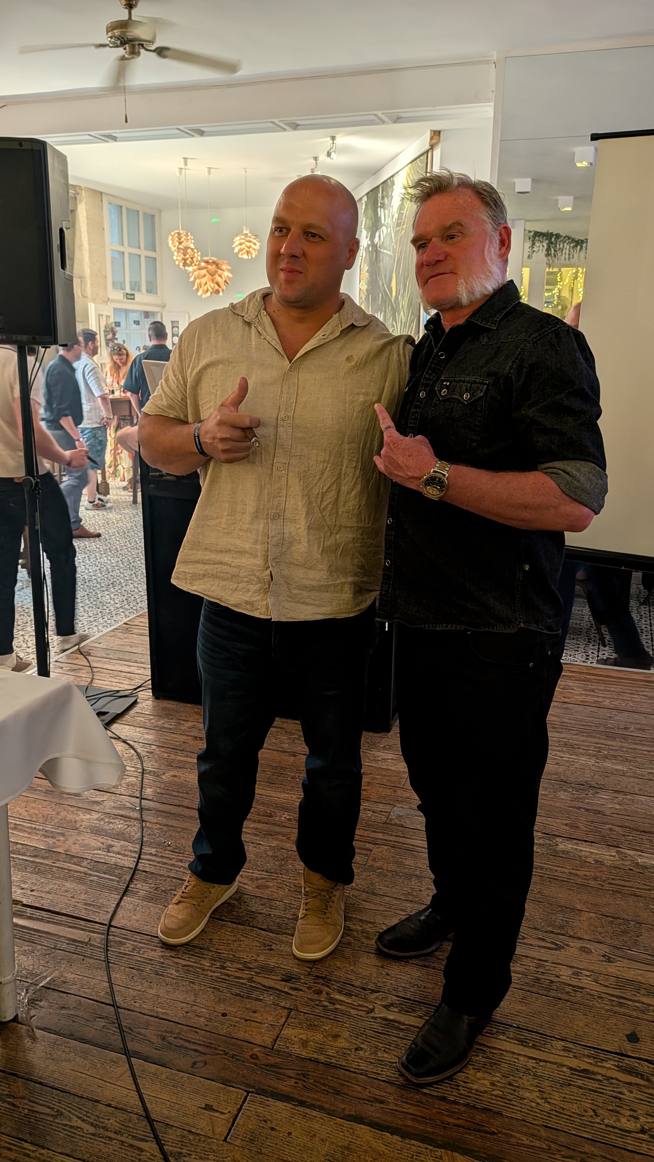 Two men posing together indoors at a social event, with people and decorations visible in the background.