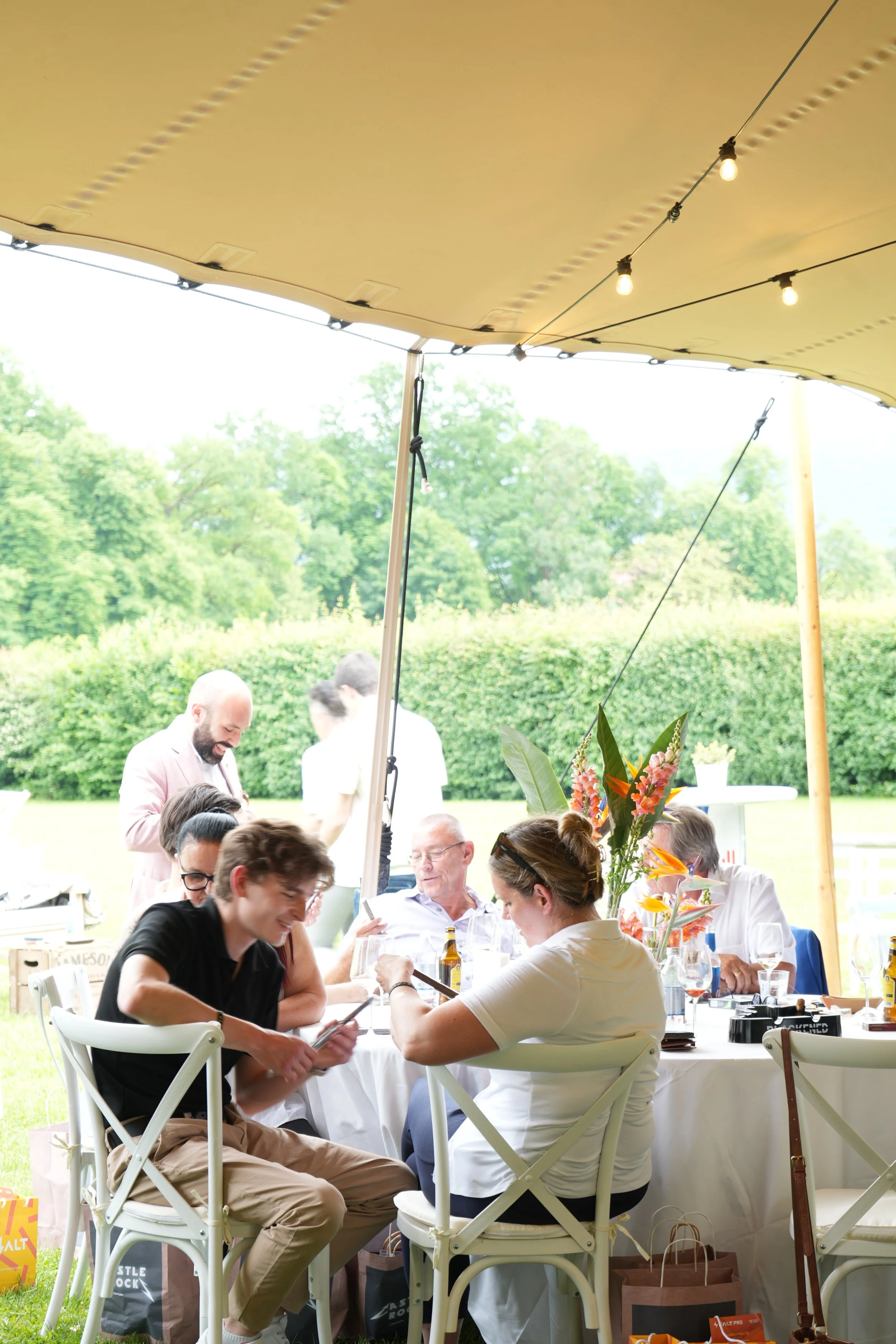 People gathered around a table under a canopy at an outdoor event, with greenery in the background.