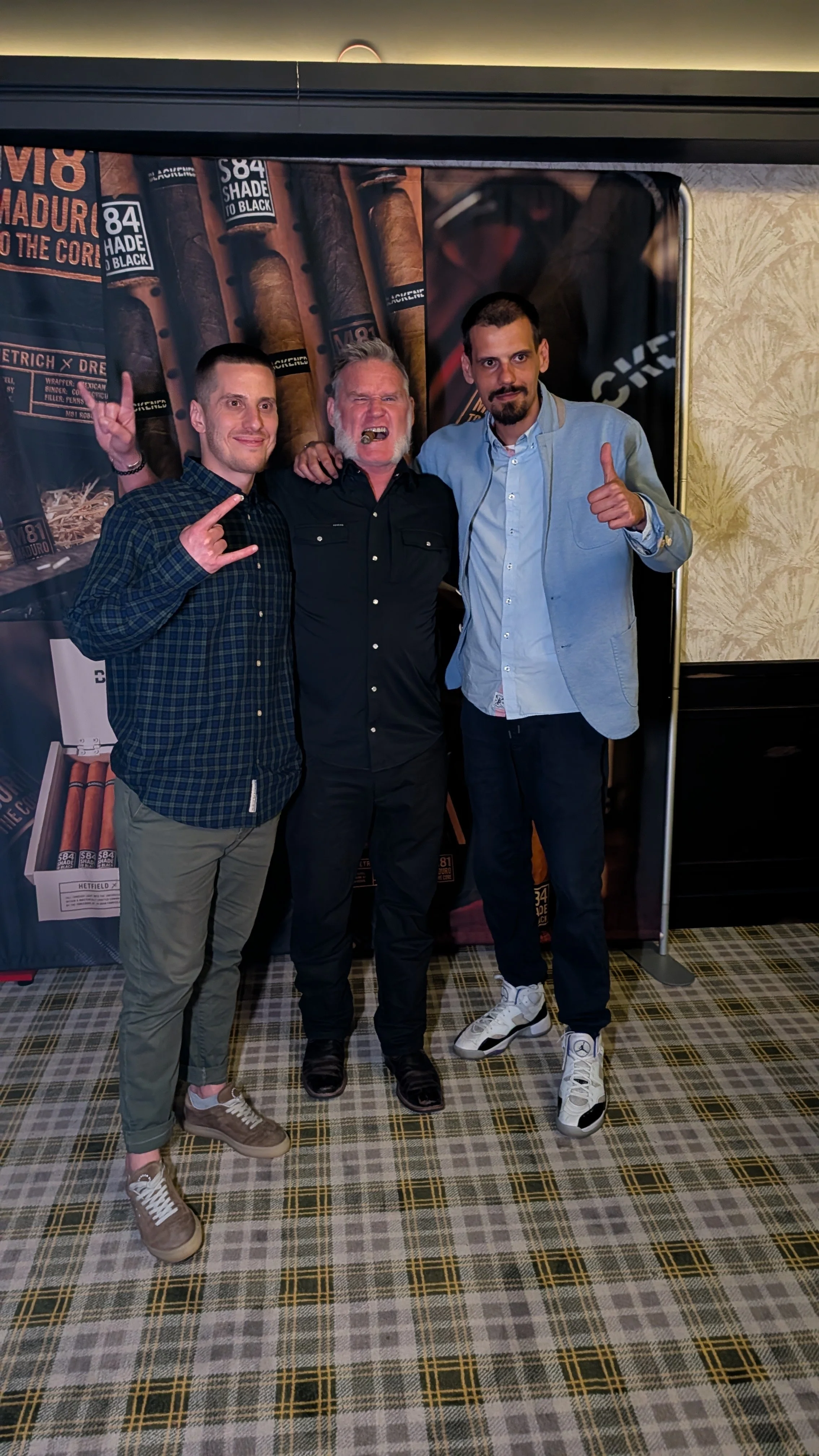 Three men standing together at a cigar event, smiling, with a backdrop of cigars and cigar branding.