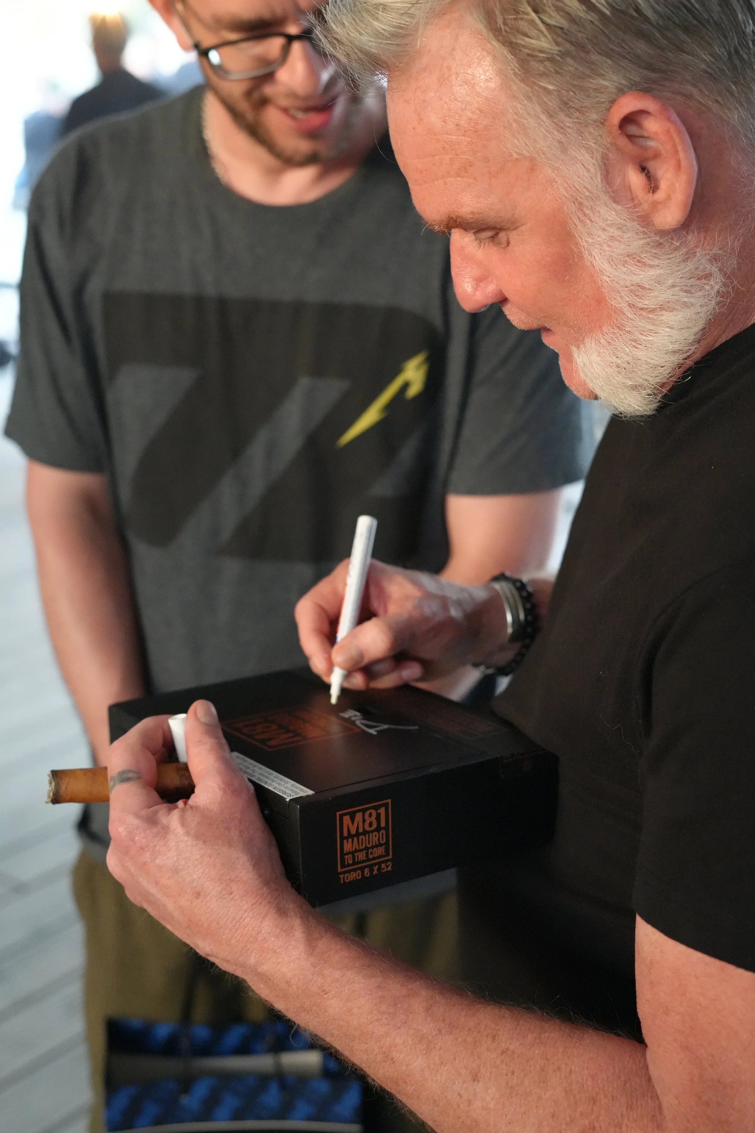 Two men are engaged in a cigarette exchange, with one man holding a box of cigars and the other signing something, indicating a transaction or signing event.