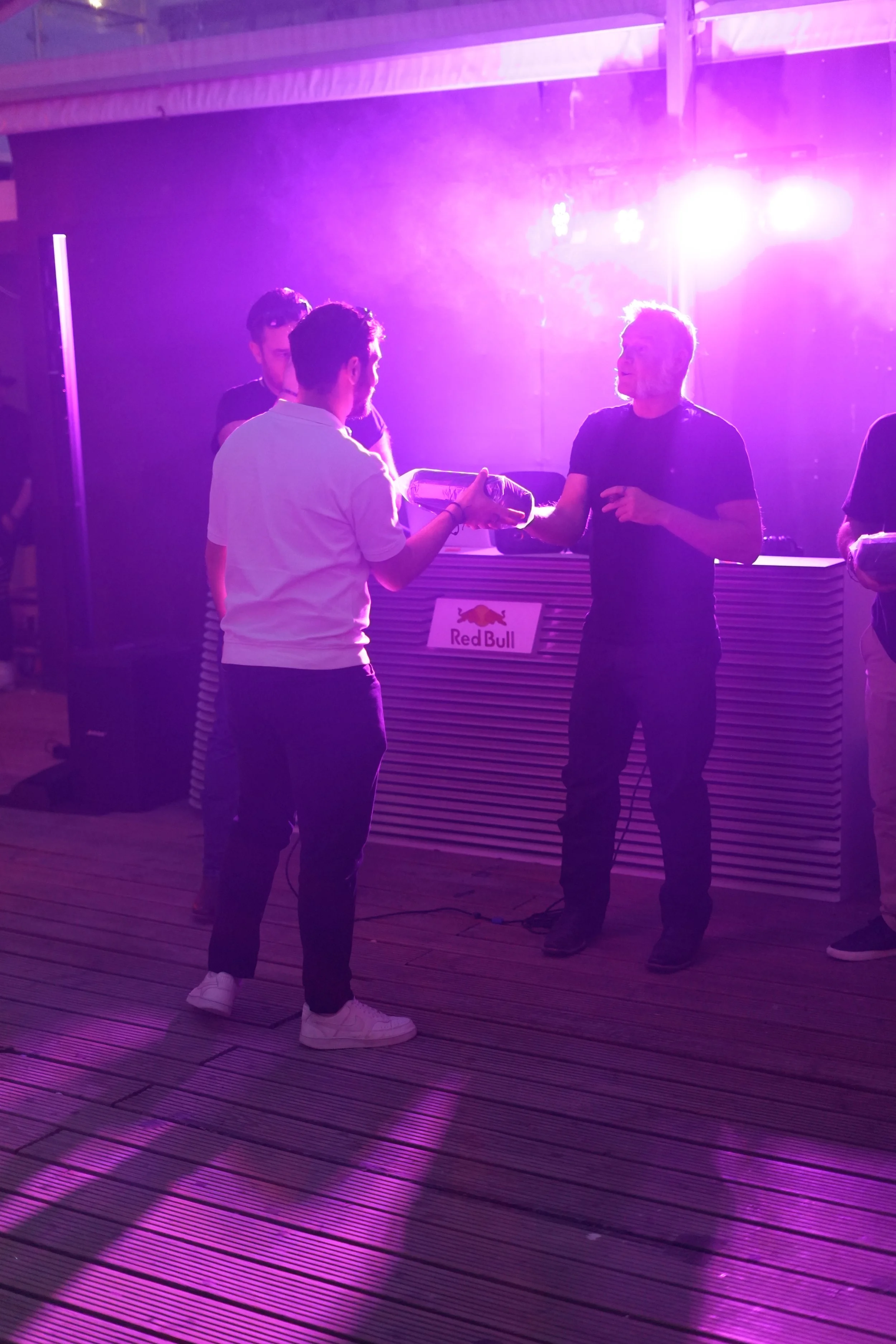 People at a bar or club with purple lighting, one person handing a bottle to another, with a Red Bull sign in the background.