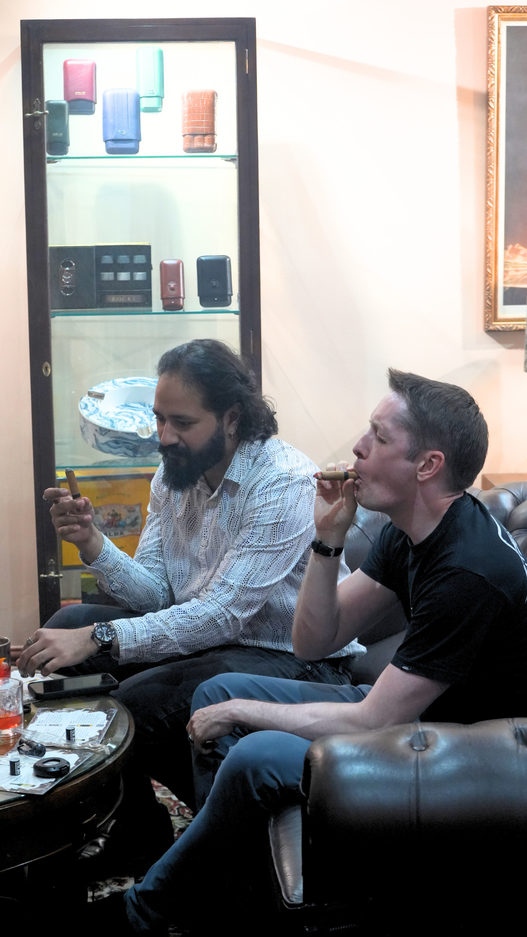 Two men sitting on a leather couch smoking cigars and having drinks in a cozy lounge room.