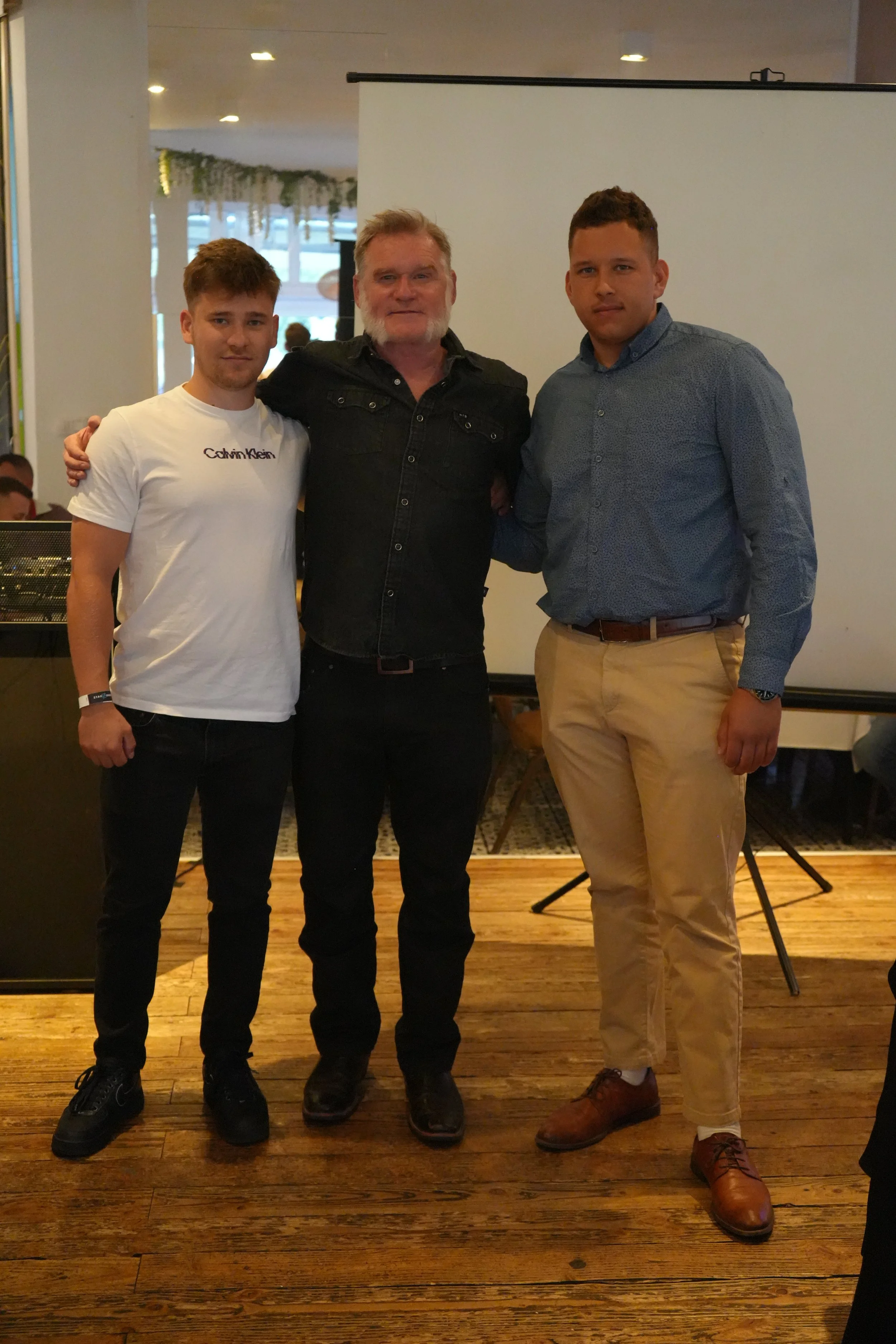 Three men stand together in an indoor setting, posing for a photo. The man on the left is wearing a white t-shirt with text, black pants, and black shoes. The man in the middle has gray hair and beard, wearing a black jacket and black pants. The man 