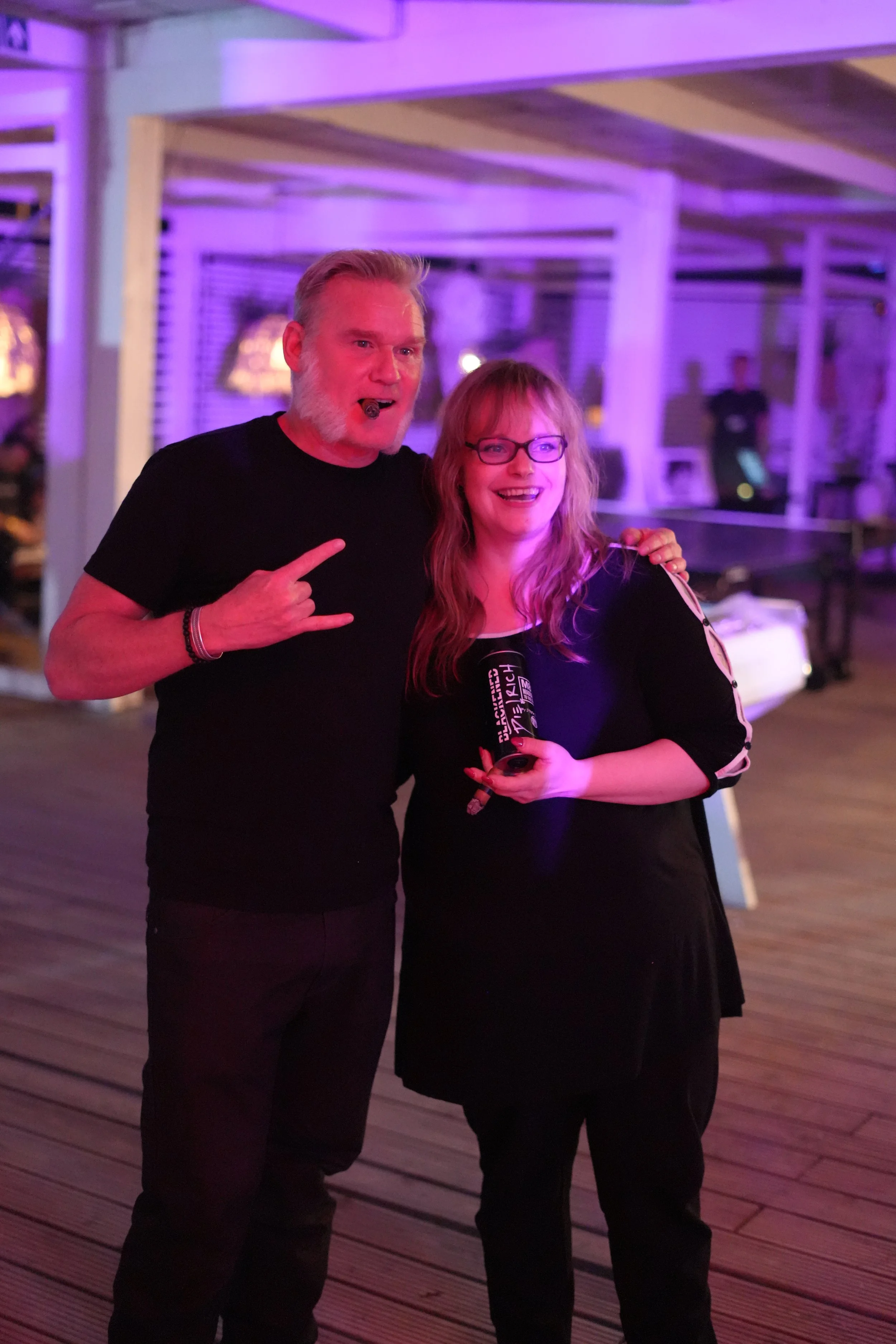 Two people standing together under purple lighting, smiling, with a man pointing at a woman who is holding a microphone and a phone. The background has a wooden floor and a blurred interior setting.