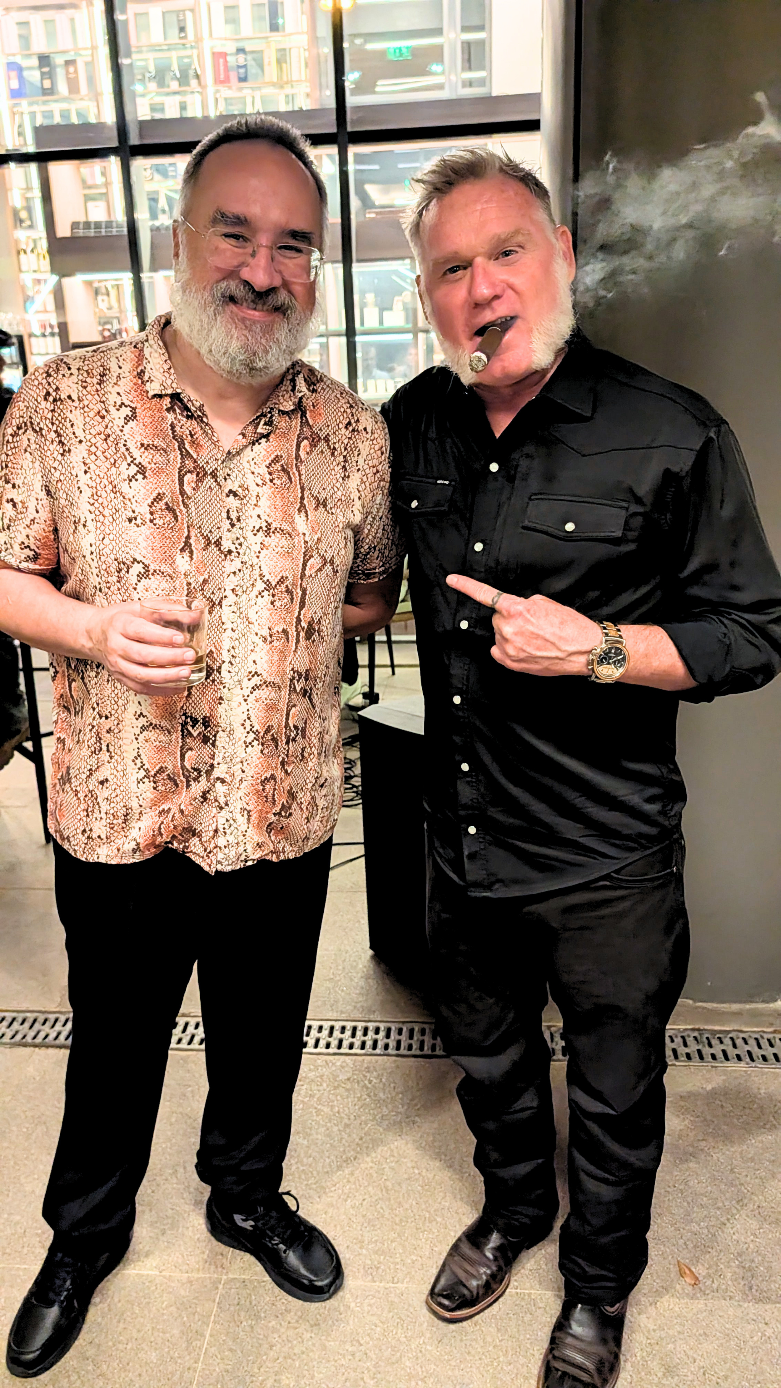 Two men standing together indoors, one wearing a snake-print shirt and glasses holding a drink, the other wearing a black shirt, smoking a cigar, and pointing at the first man, with a cityscape visible through large windows behind them.