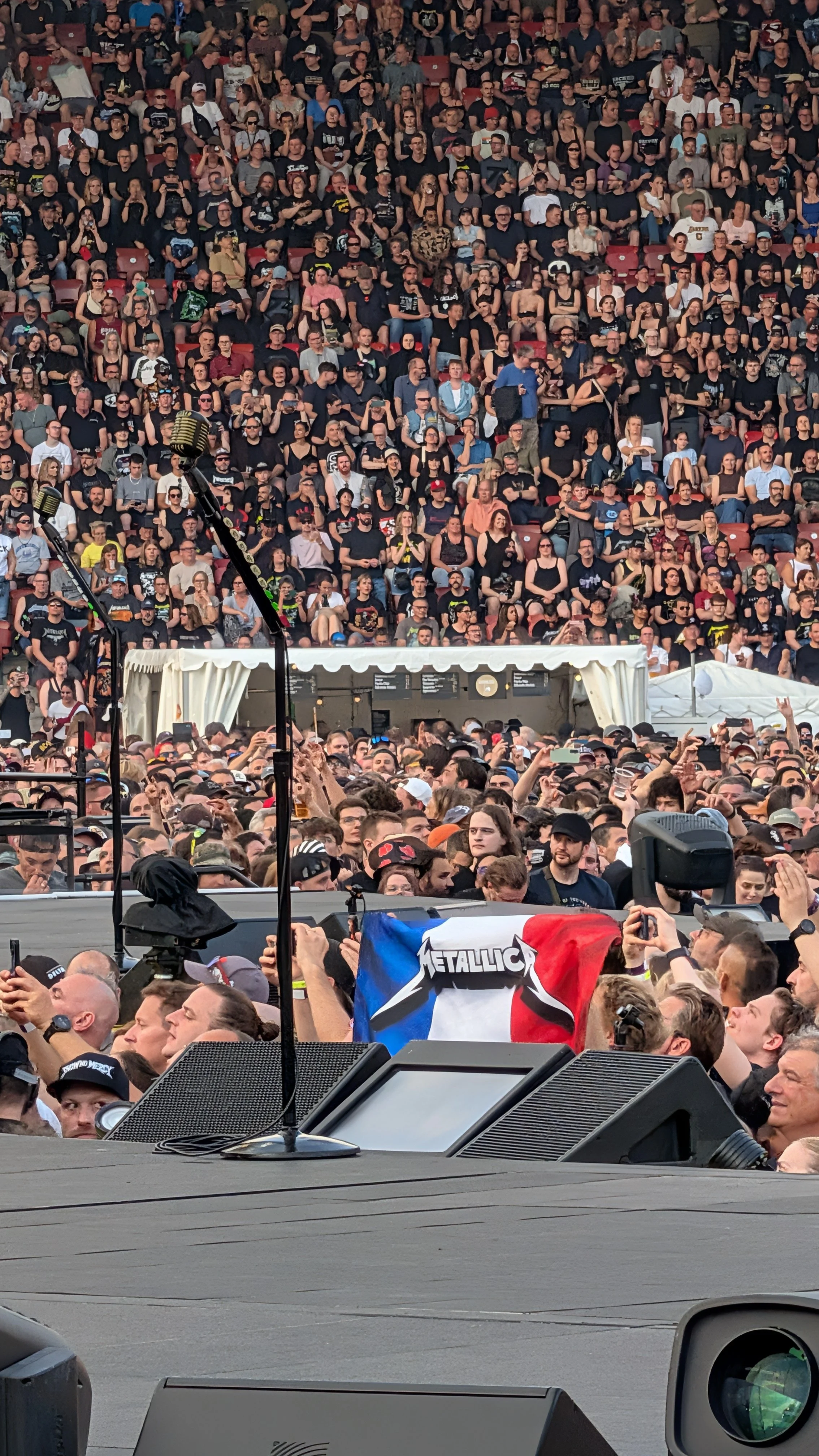 Crowd at a Metallica concert, with many people standing and sitting, some taking photos, and a flag with the Metallica band logo is visible among the crowd.