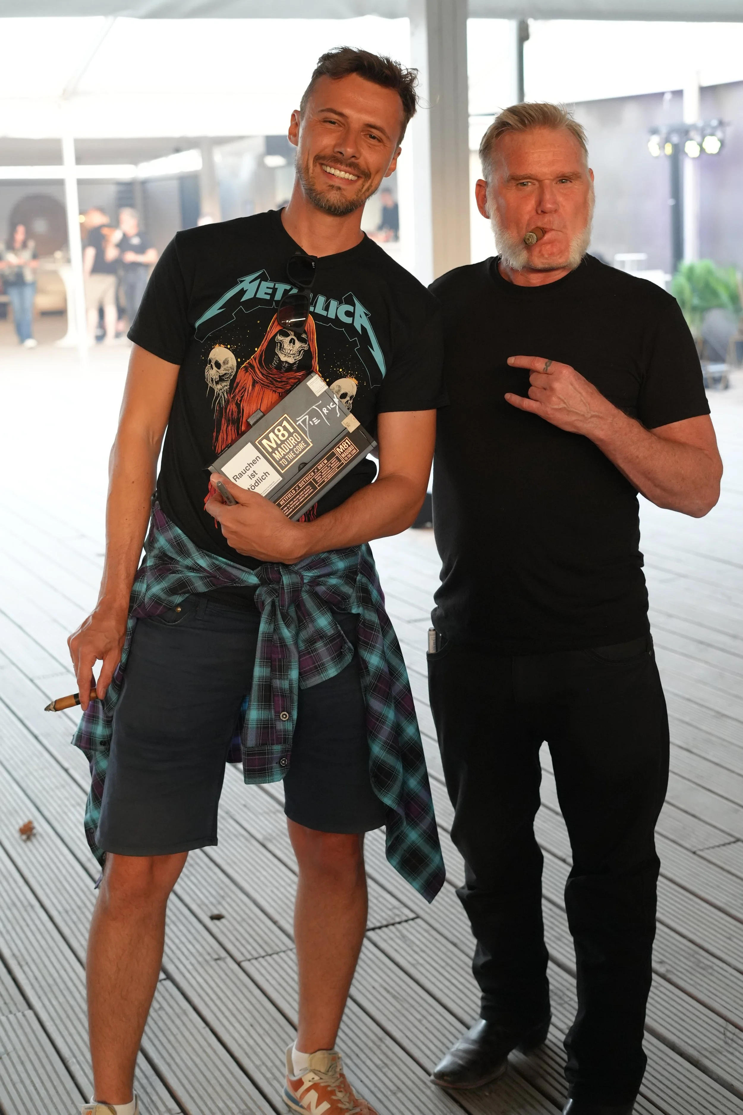 Two men standing together indoors, smiling, and posing for the photo. One man is wearing a black Metallica t-shirt, plaid shirt tied around his waist, shorts, and sneakers, holding a small package. The other man is dressed in black, with gray hair, s