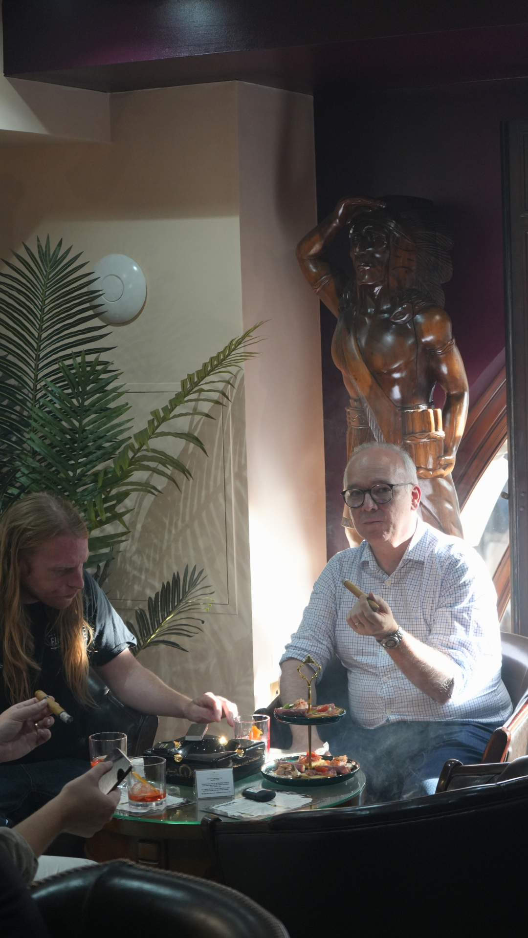 People sitting at a table with plates of food and drinks, in a restaurant with a large wooden sculpture of a muscular man with a headdress in the background, and tropical plants on the wall.