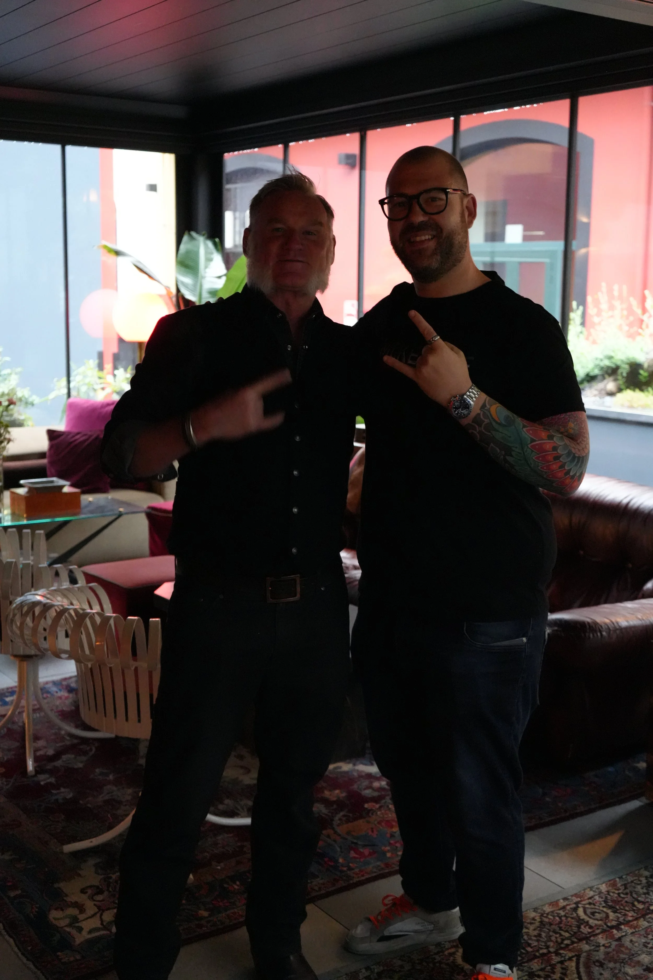 Two men standing together indoors, silhouetted against a window with colorful exterior, one wearing glasses and a tattooed arm, the other with a beard. They are smiling and making hand gestures.