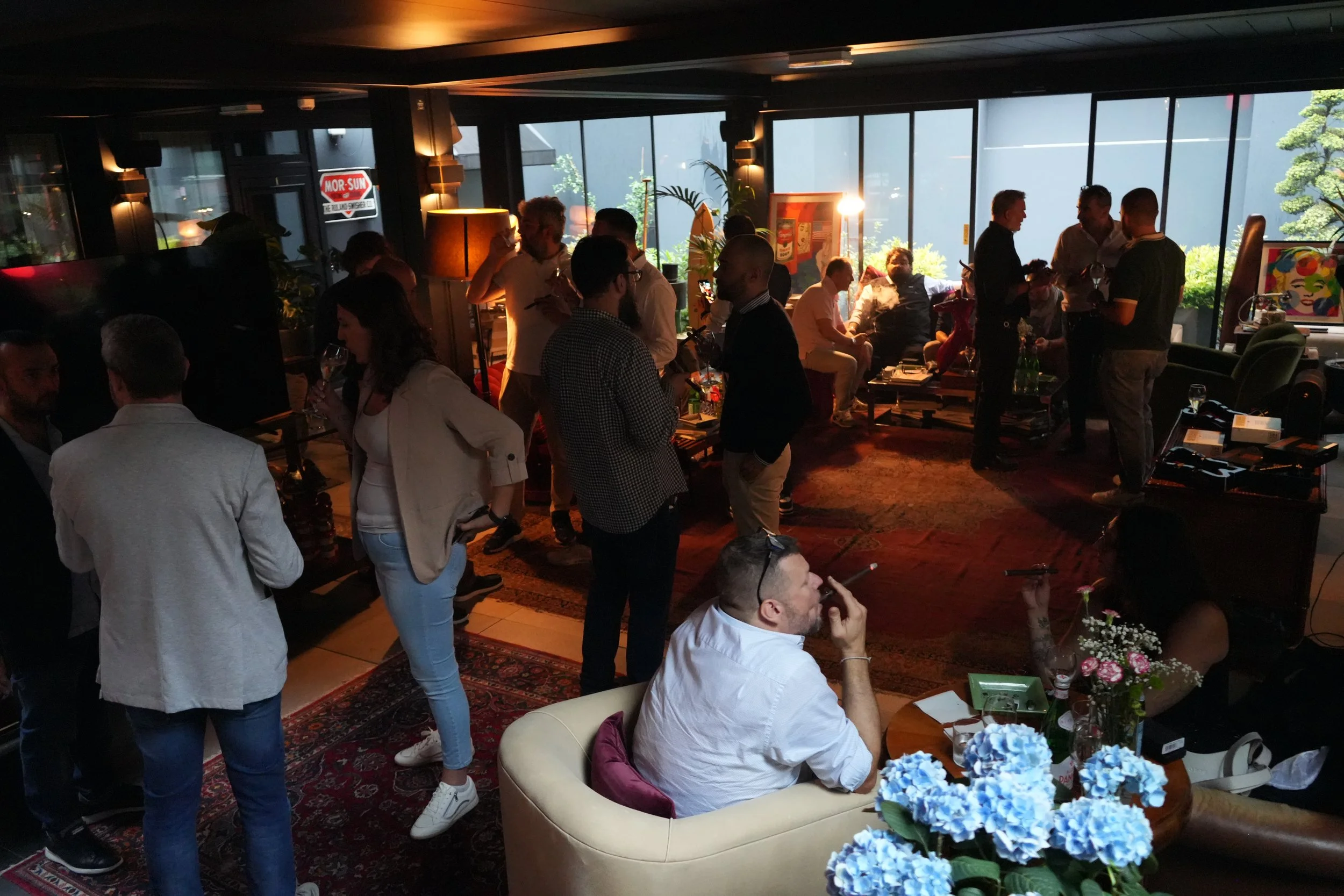 People gathered in a cozy, dimly lit room with large windows showing greenery outside. Many individuals are engaging in conversations, with some holding drinks. The room has a warm ambiance with lamps and decorative artwork. A man is seated on a crea
