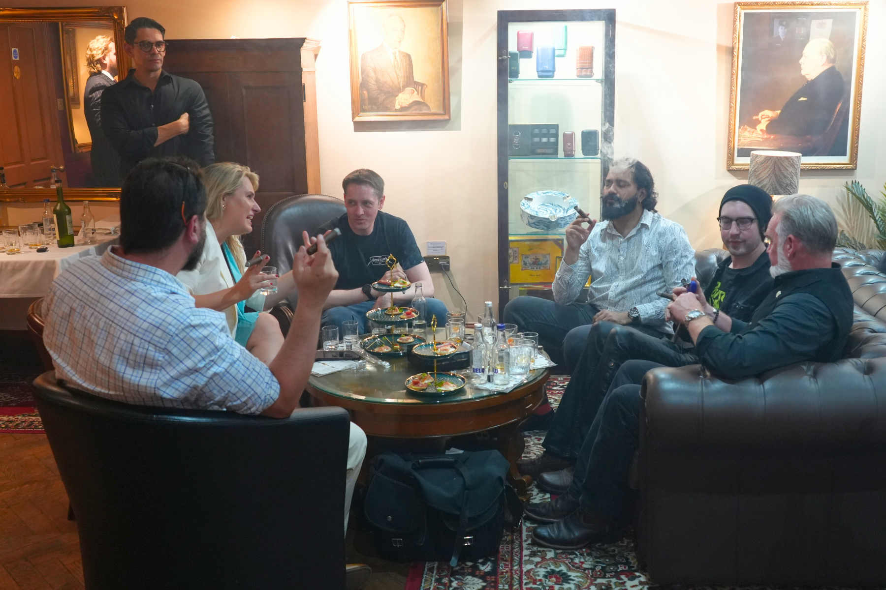 Group of people sitting around a coffee table in a living room, socializing and smoking. There are paintings on the wall, glass bottles, and small plates of food on the table.