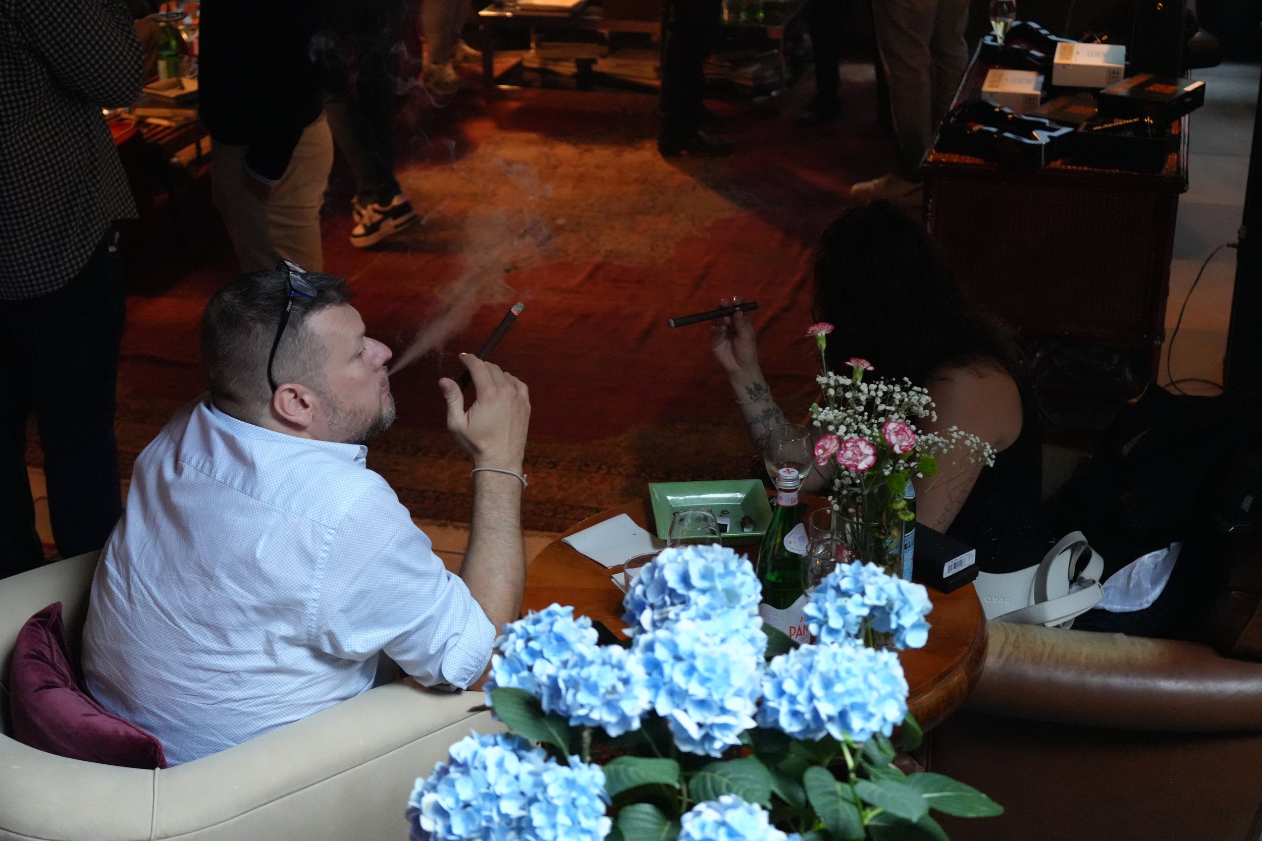 A man with short hair and a beard sits at a table smoking a cigar, with blue hydrangea flowers in the foreground. A woman with dark hair and tattoos on her arm is also sitting nearby with a flower tucked behind her ear. The scene appears to be a soci