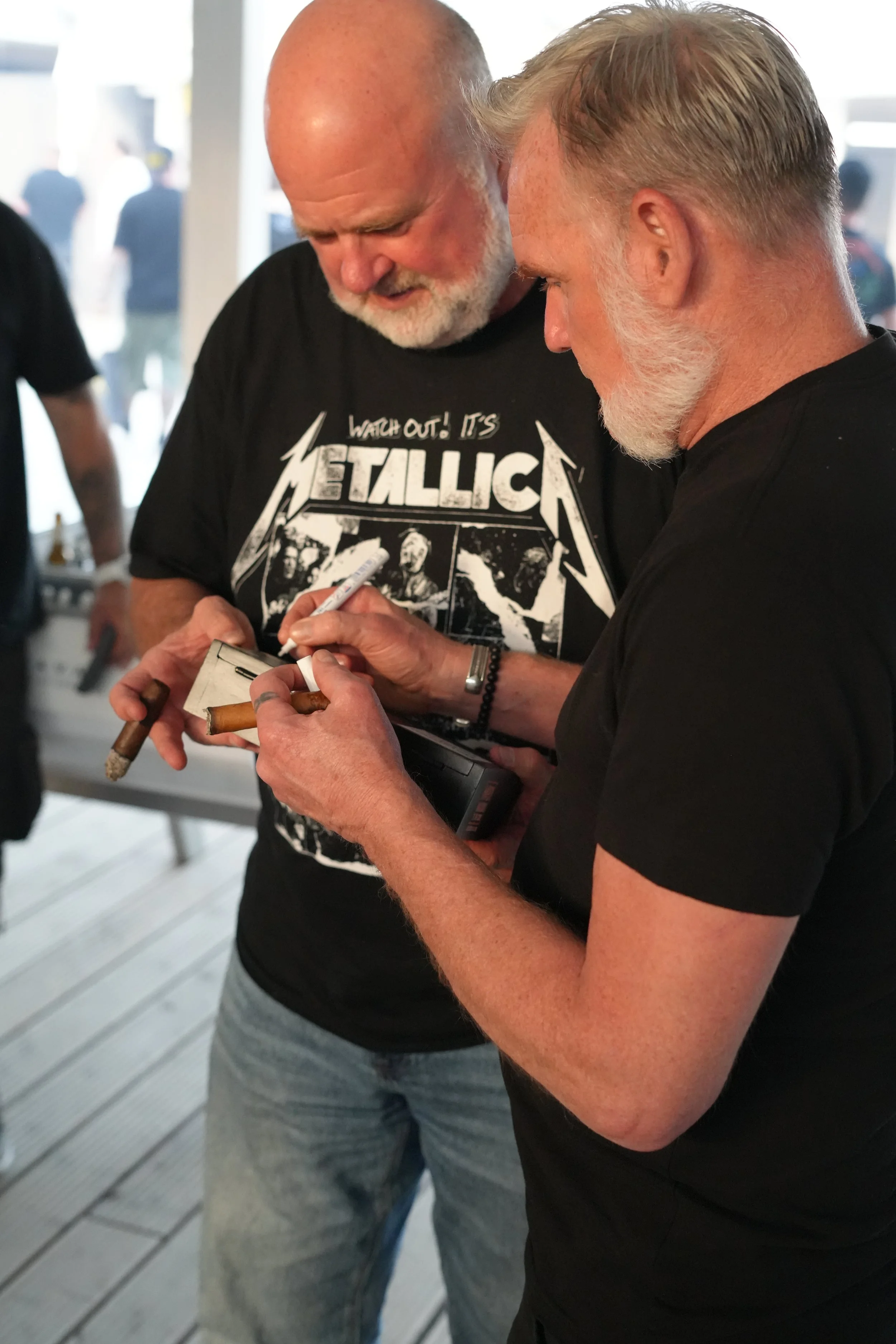 Two men, one with a beard and wearing a black Metallica T-shirt, and the other with a lighter beard and a black shirt, are signing an item together. They are standing close, engaged in the activity, with a blurred background consisting of people and 