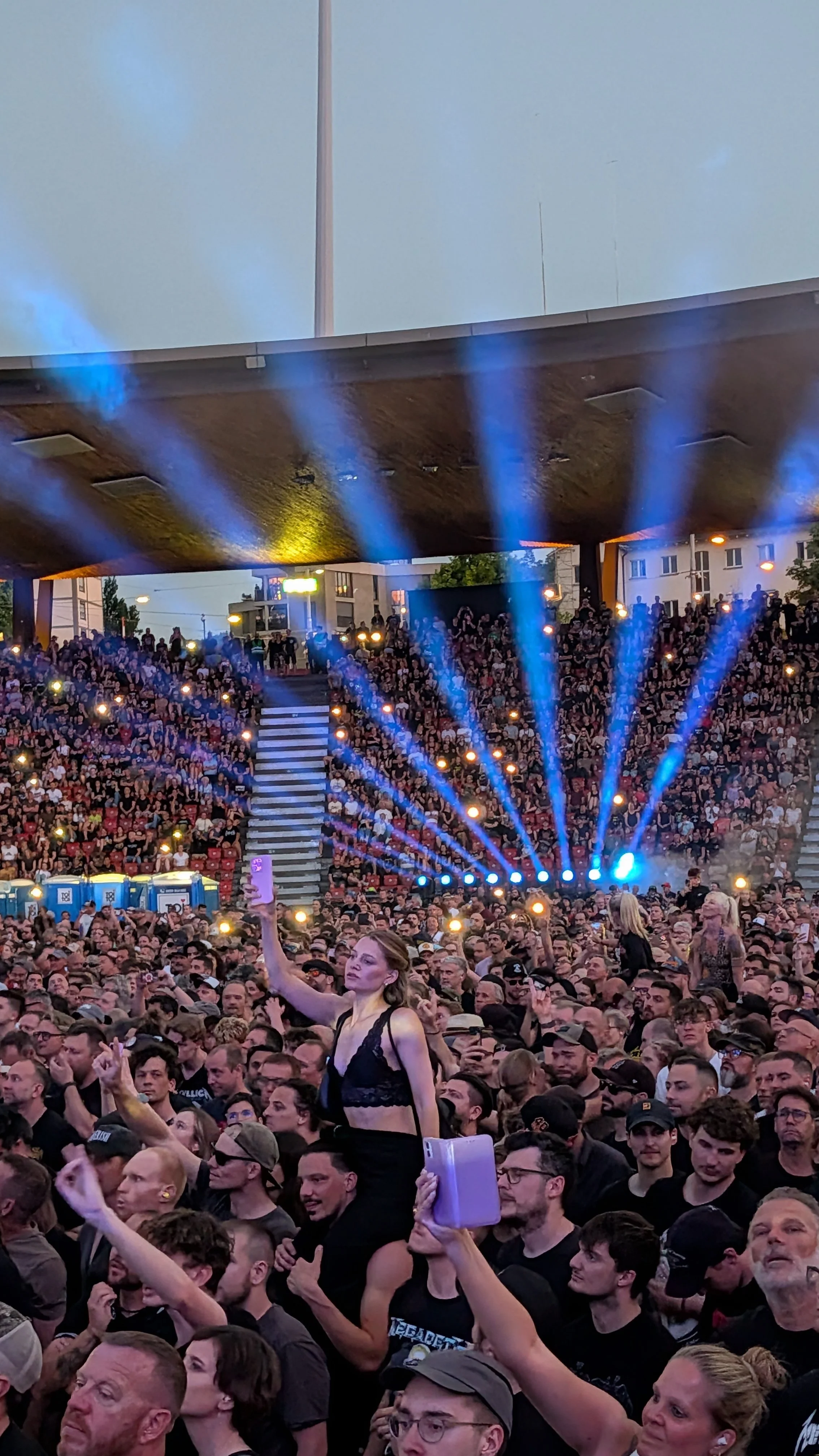 Crowd attending a Metallica concert with a stage and blue lights, and a woman in the foreground taking a selfie with her phone.