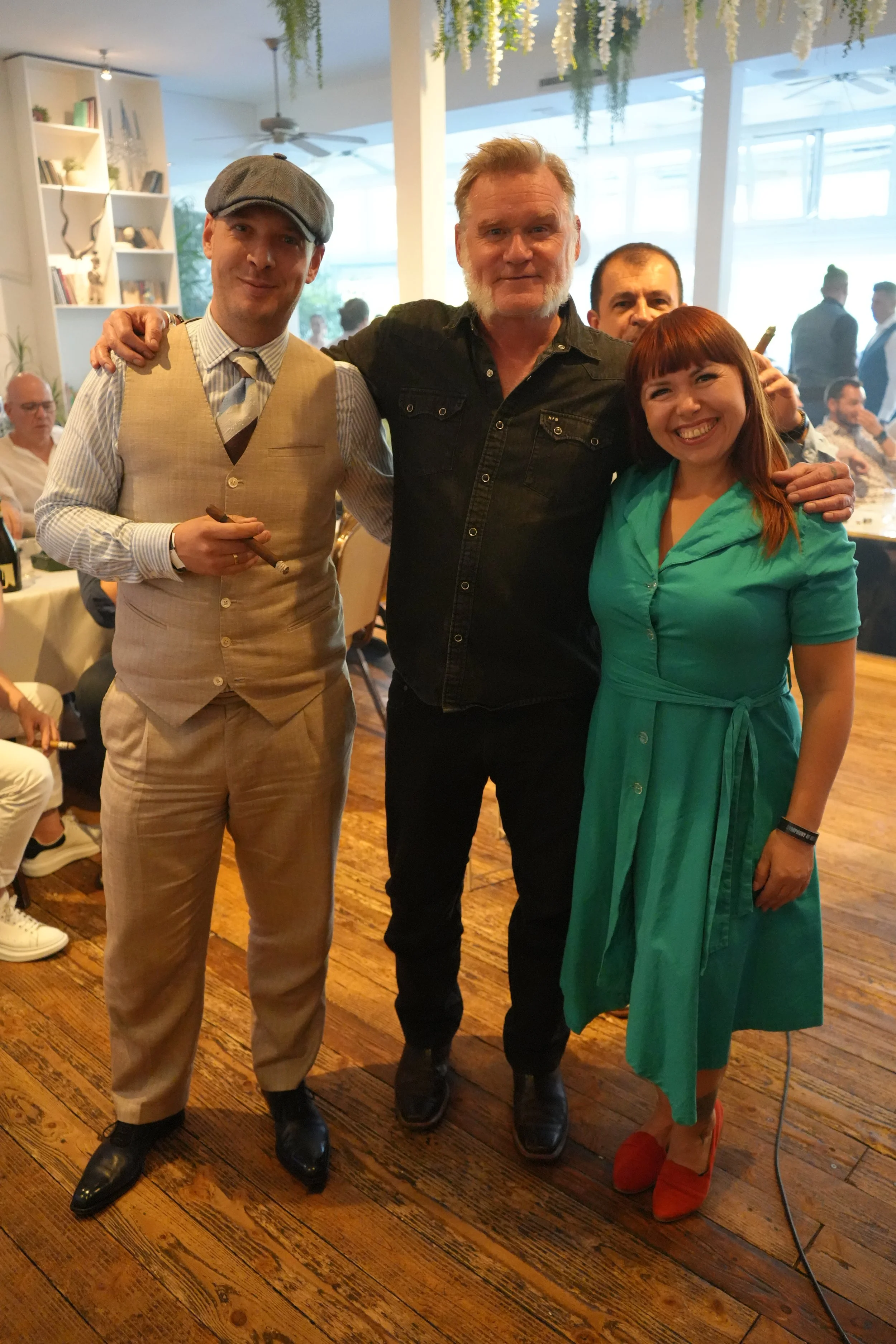 Four people are smiling and posing together at a social gathering indoors. One man on the left is dressed in vintage style with a beige vest, striped shirt, and flat cap, holding a cigar. The man in the center has a beard and is wearing a black shirt