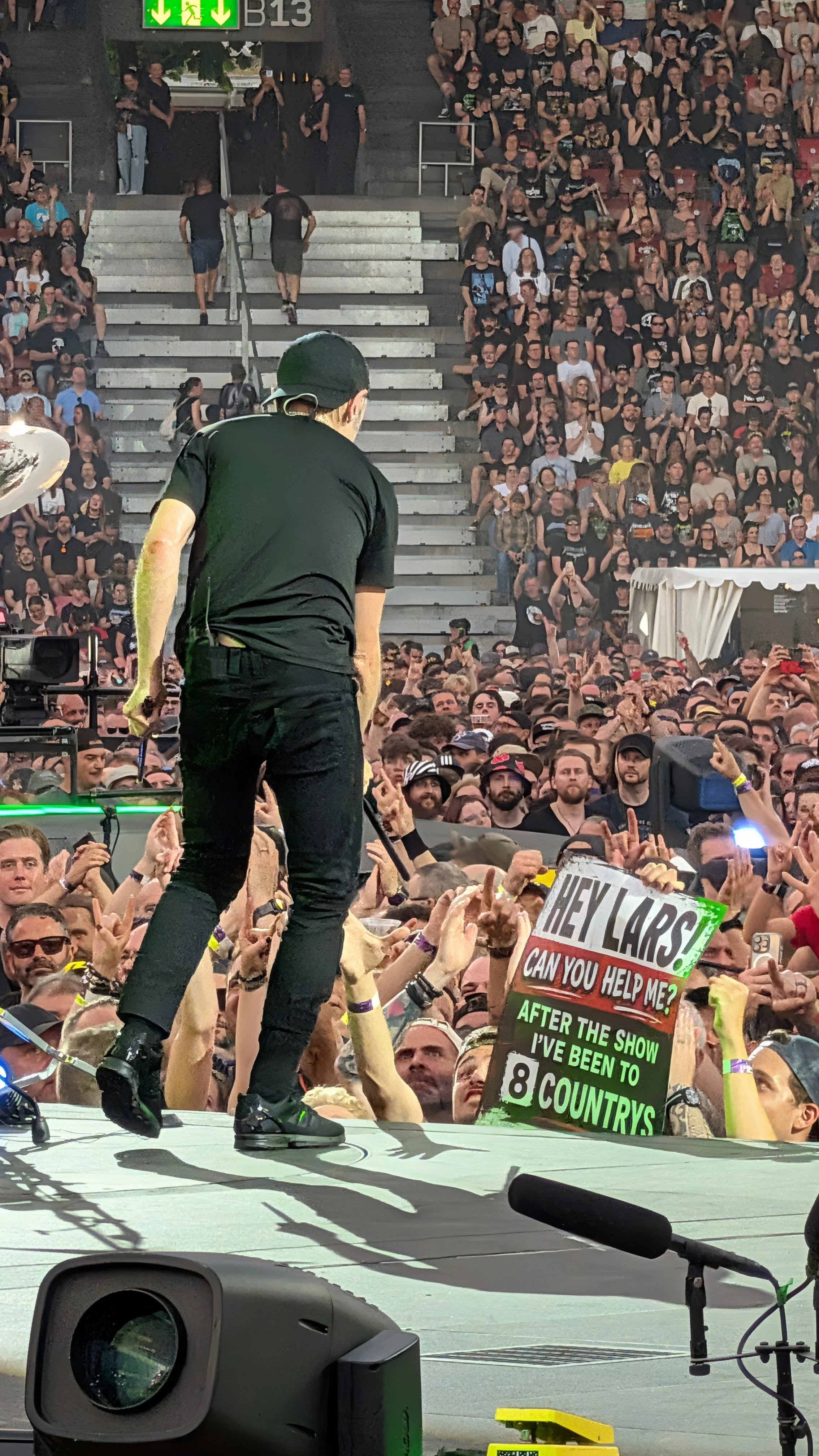 Crowd at a Metallica concert or festival with Lars Ulrich on stage, and many people holding up signs and taking photos