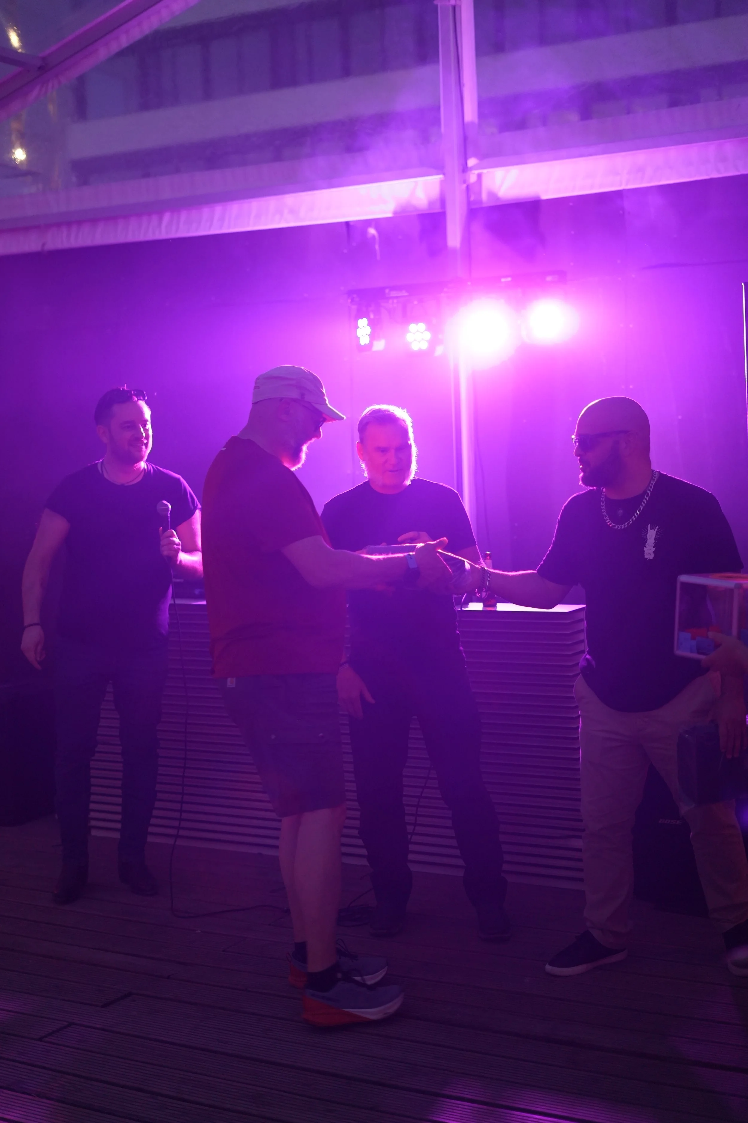 Four men are standing under purple stage lights in a dimly lit indoor setting. One man is holding a microphone, another is handing over a small object, while the others observe or smile. The atmosphere suggests a casual event or celebration.