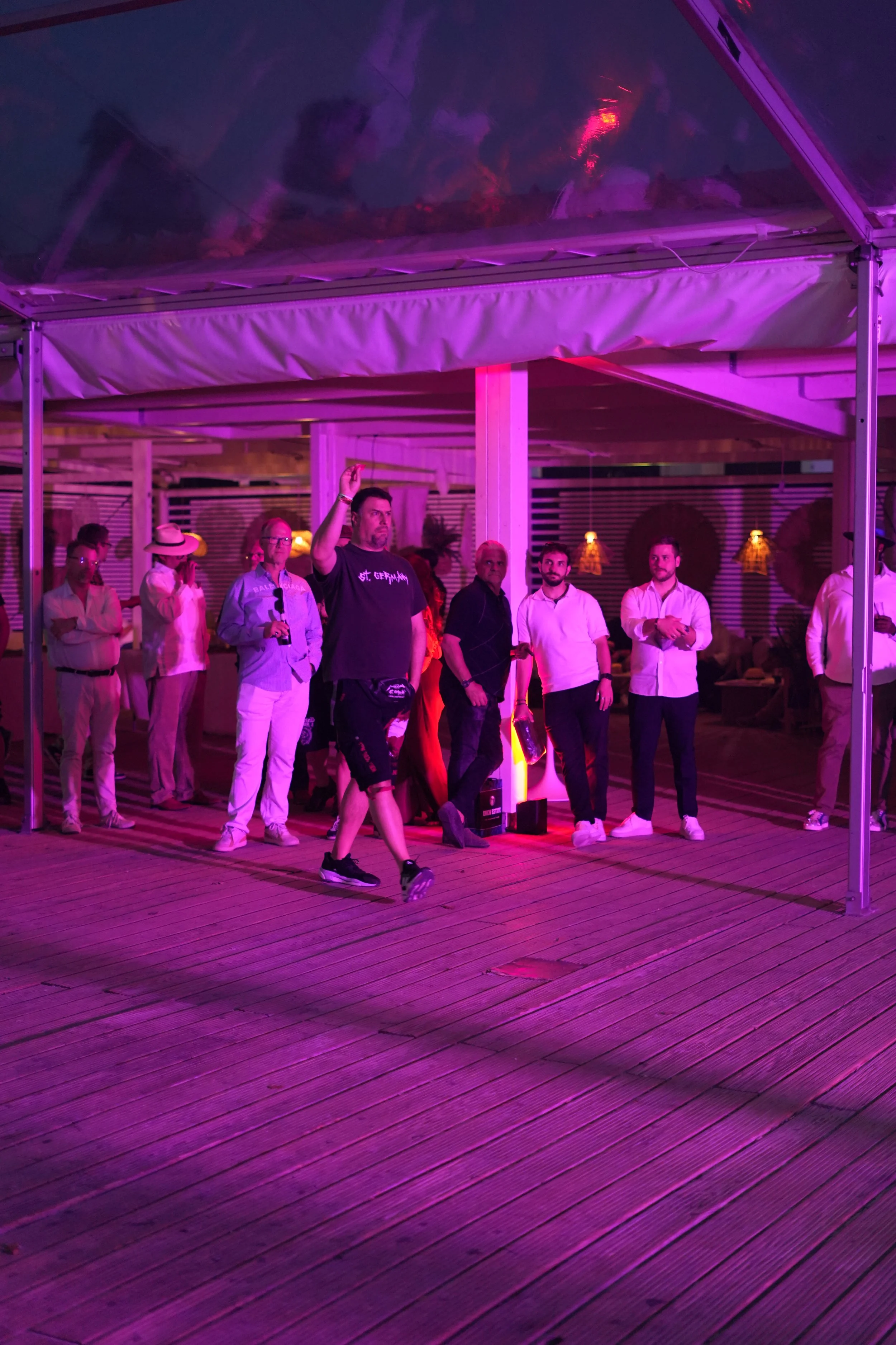 People gathered under a purple-lit canopy on a wooden deck, with some dressed in white and one person in a black T-shirt raising a hand.
