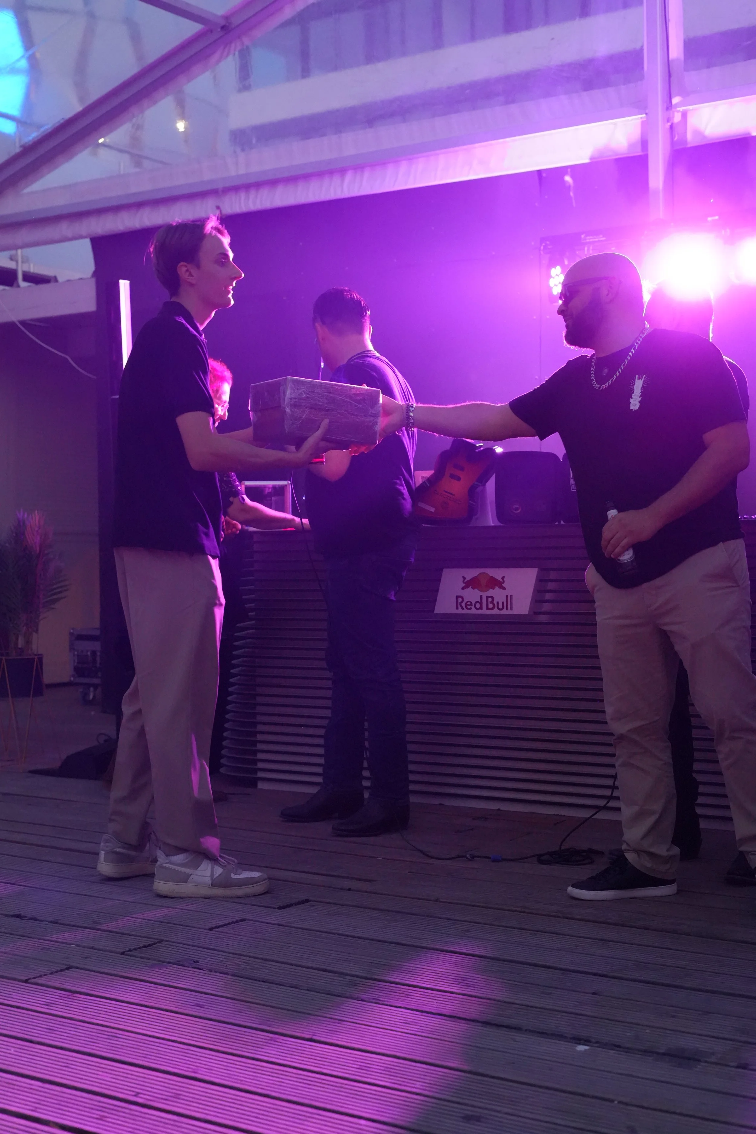 Two men exchanging a wrapped gift at an indoor event with purple lighting, Red Bull logo visible on a panel, and DJ equipment in the background.