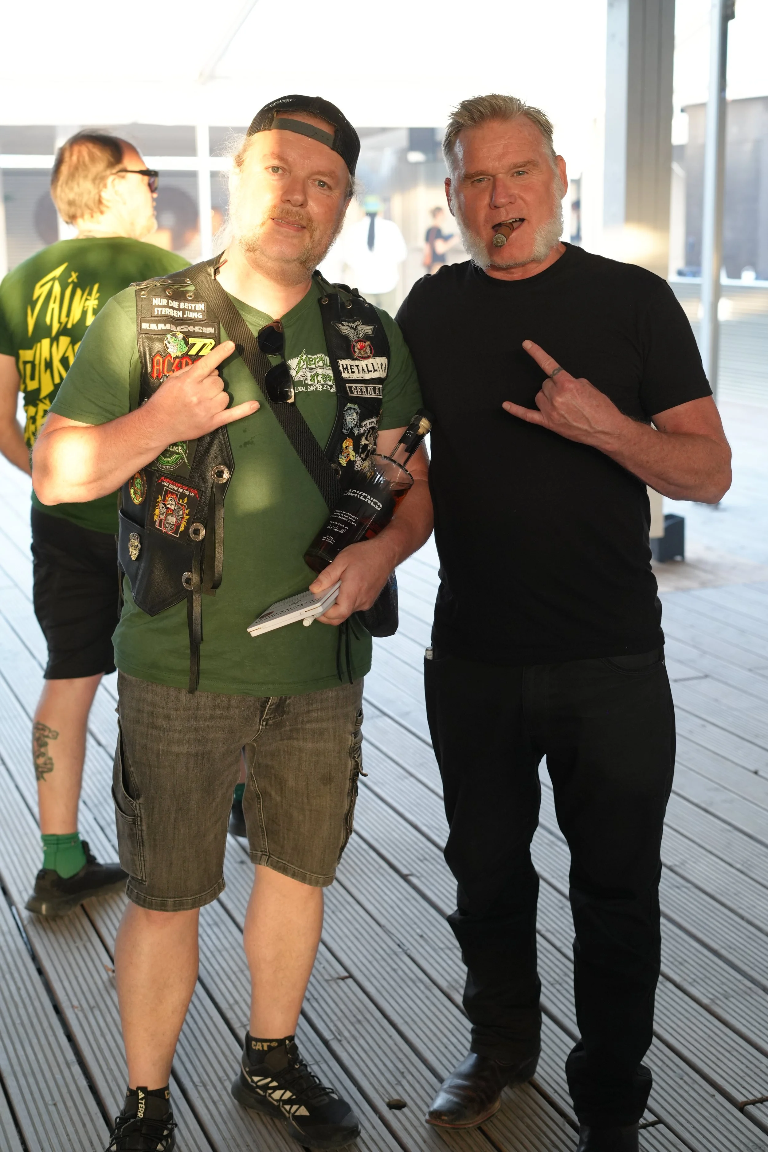 Two men standing on a wooden deck, making rock signs with their hands, one wearing a black shirt and cigar and the other wearing a vest with patches and a backward cap, smiling at the camera.
