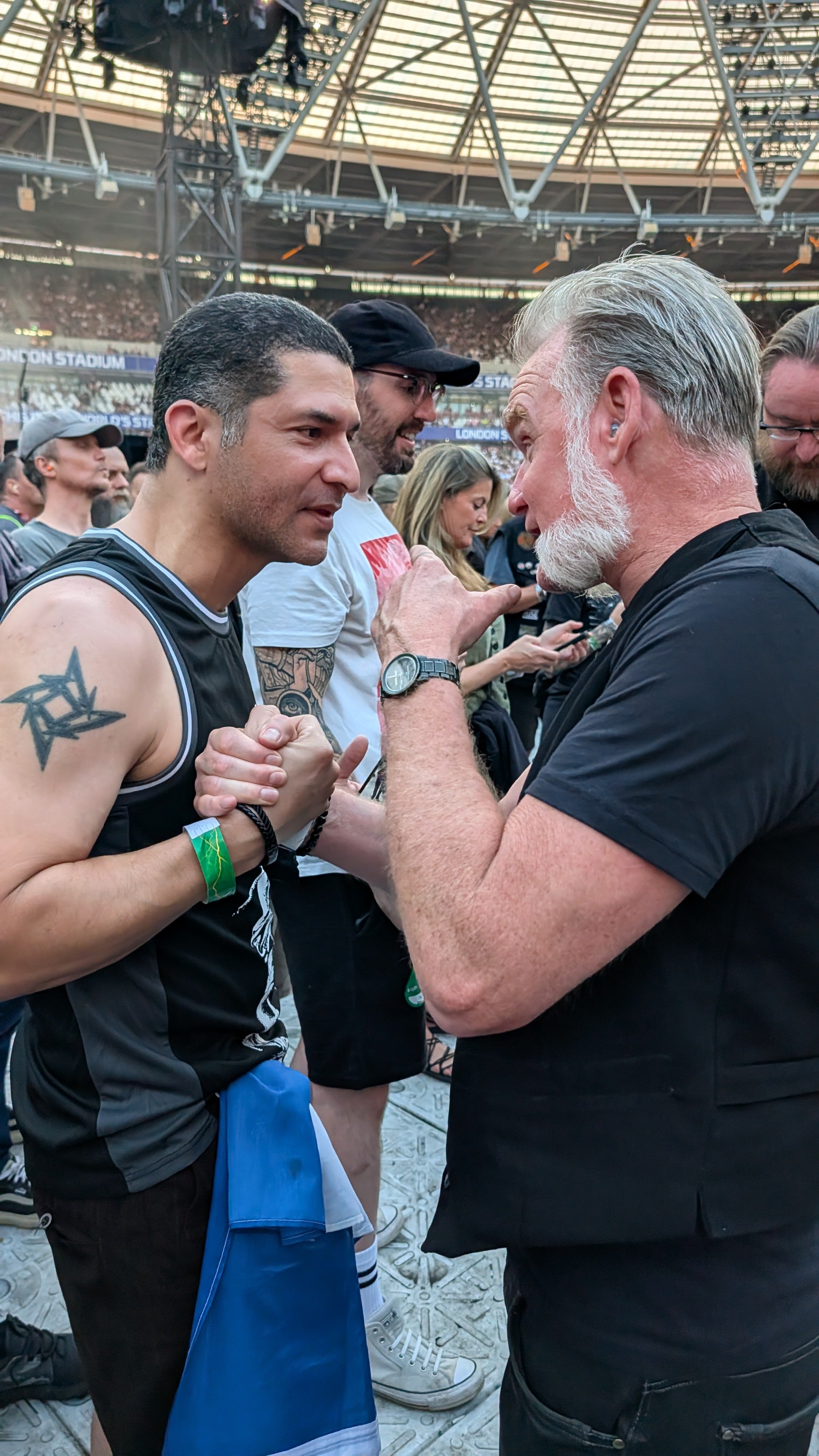 Two men are in a conversation with their hands clasped and leaning toward each other at a stadium. The man on the left is wearing a black sports jersey with a tattoo on his right shoulder, and the older man on the right has a gray beard and is wearin