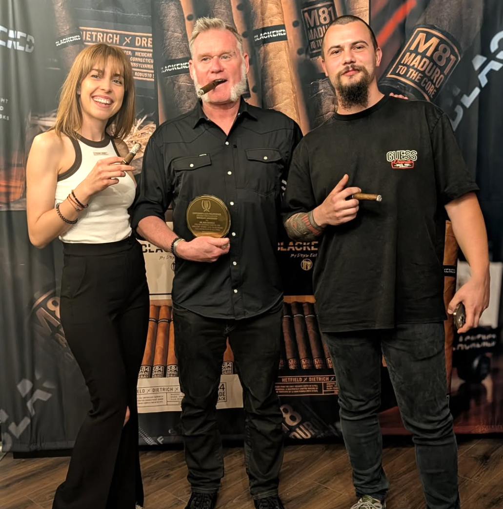Three people standing together, holding cigars, in front of a cigar-themed backdrop. The woman on the left is smiling, the man in the middle is holding a plaque and has a cigar in his mouth, and the man on the right is also smiling and holding a ciga
