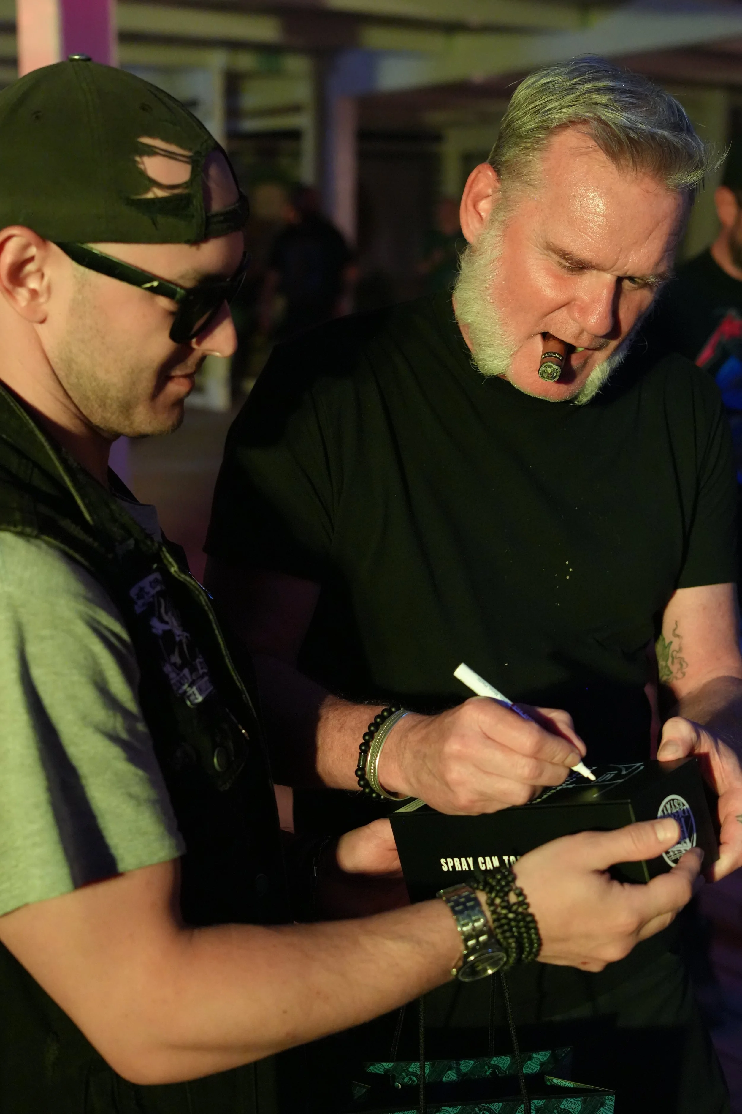 Two men are interacting at night, one with a motorcycle vest and sunglasses and the other with gray hair, holding a black box with writing, wearing a black T-shirt, and smoking a cigar. The scene appears to be at an event or gathering.
