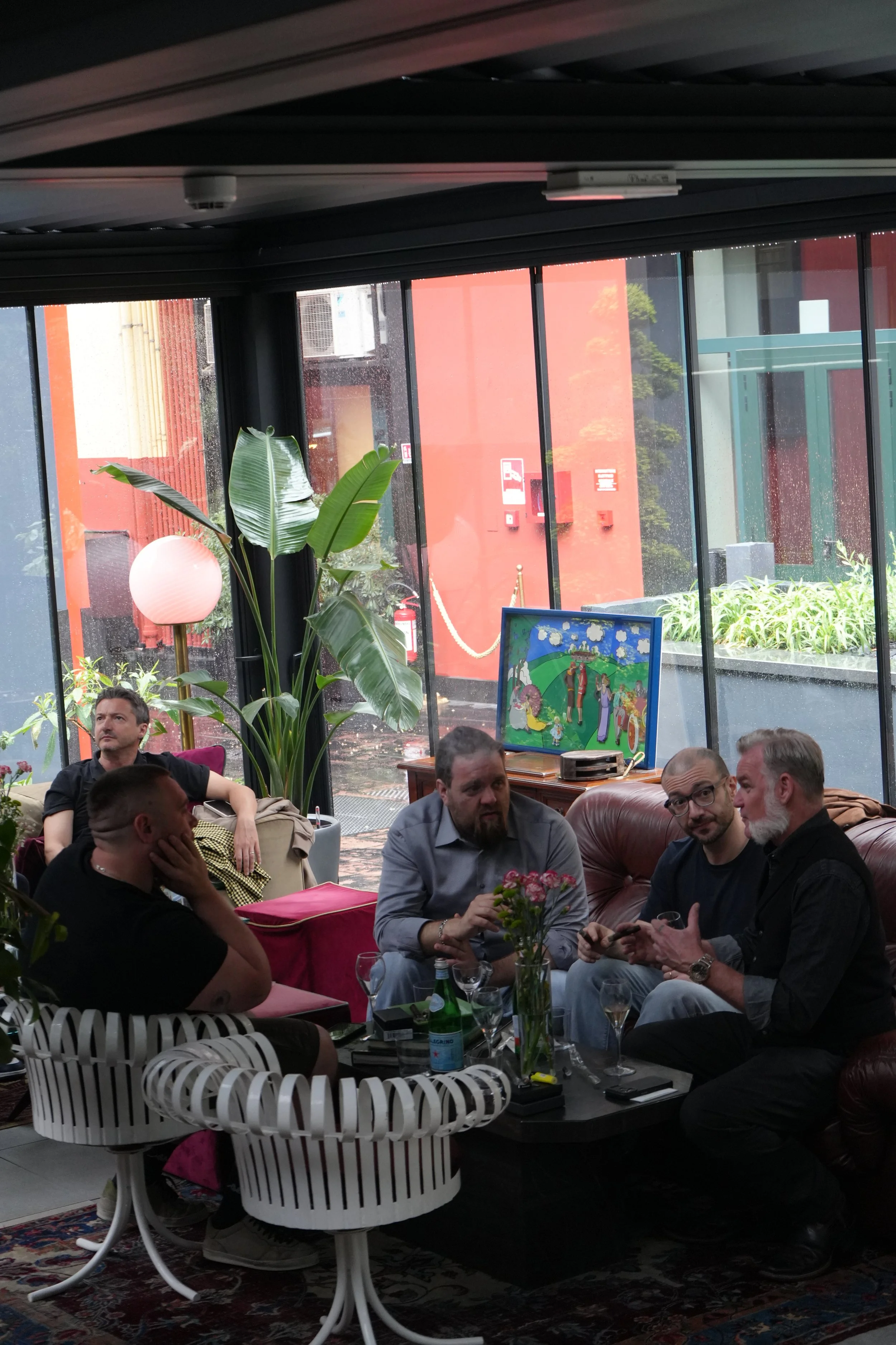 A group of people sitting inside a room with large windows, decorated with plants and artwork, having a conversation and socializing.