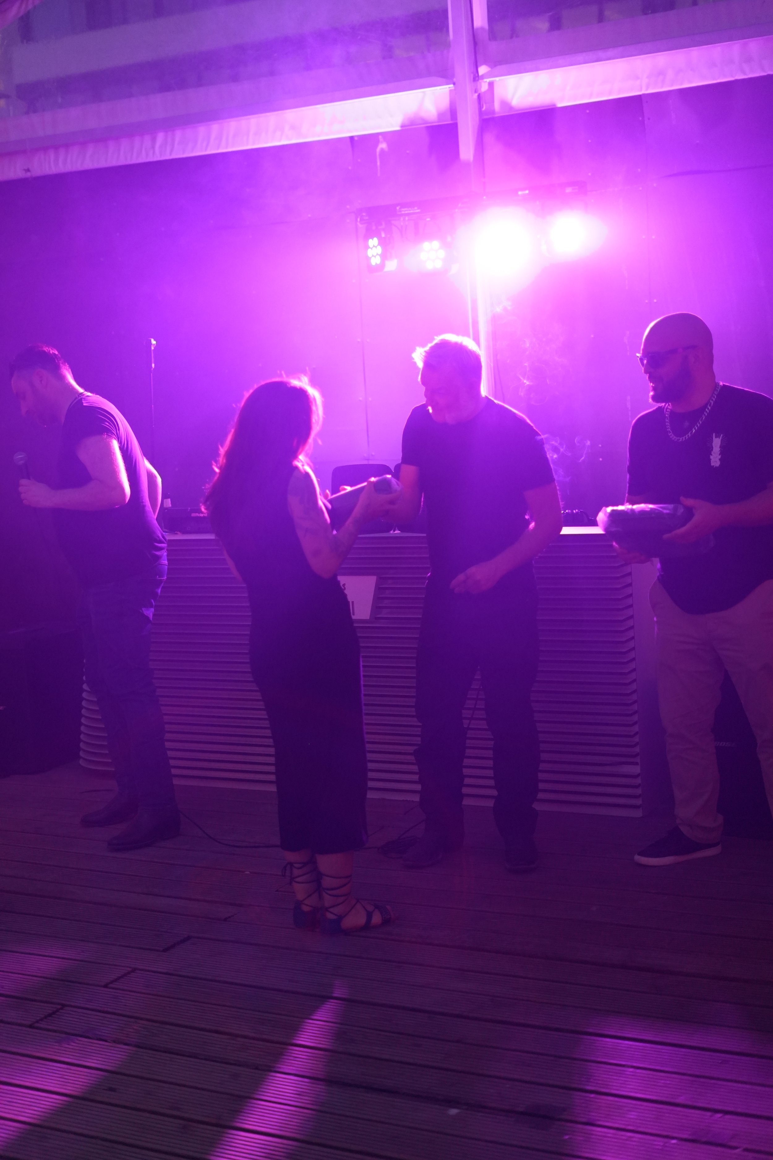 Four people standing in a club with purple lighting, socializing. One woman has tattoos and is looking at her phone, while three men are facing her, two of whom are smiling. The man on the left is holding a microphone.