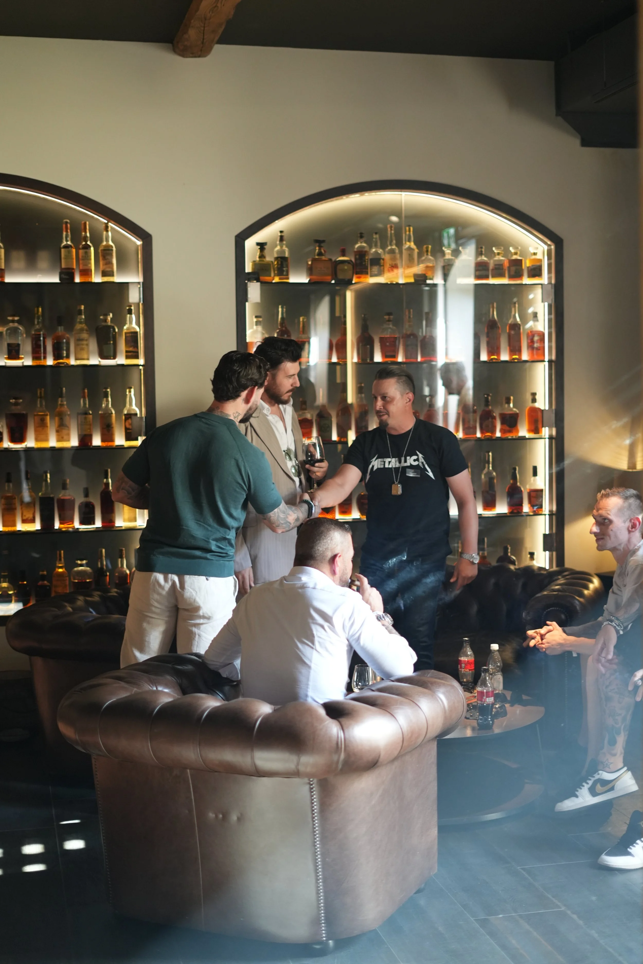 Group of people socializing in a lounge or bar with liquor bottles on illuminated shelves in the background.