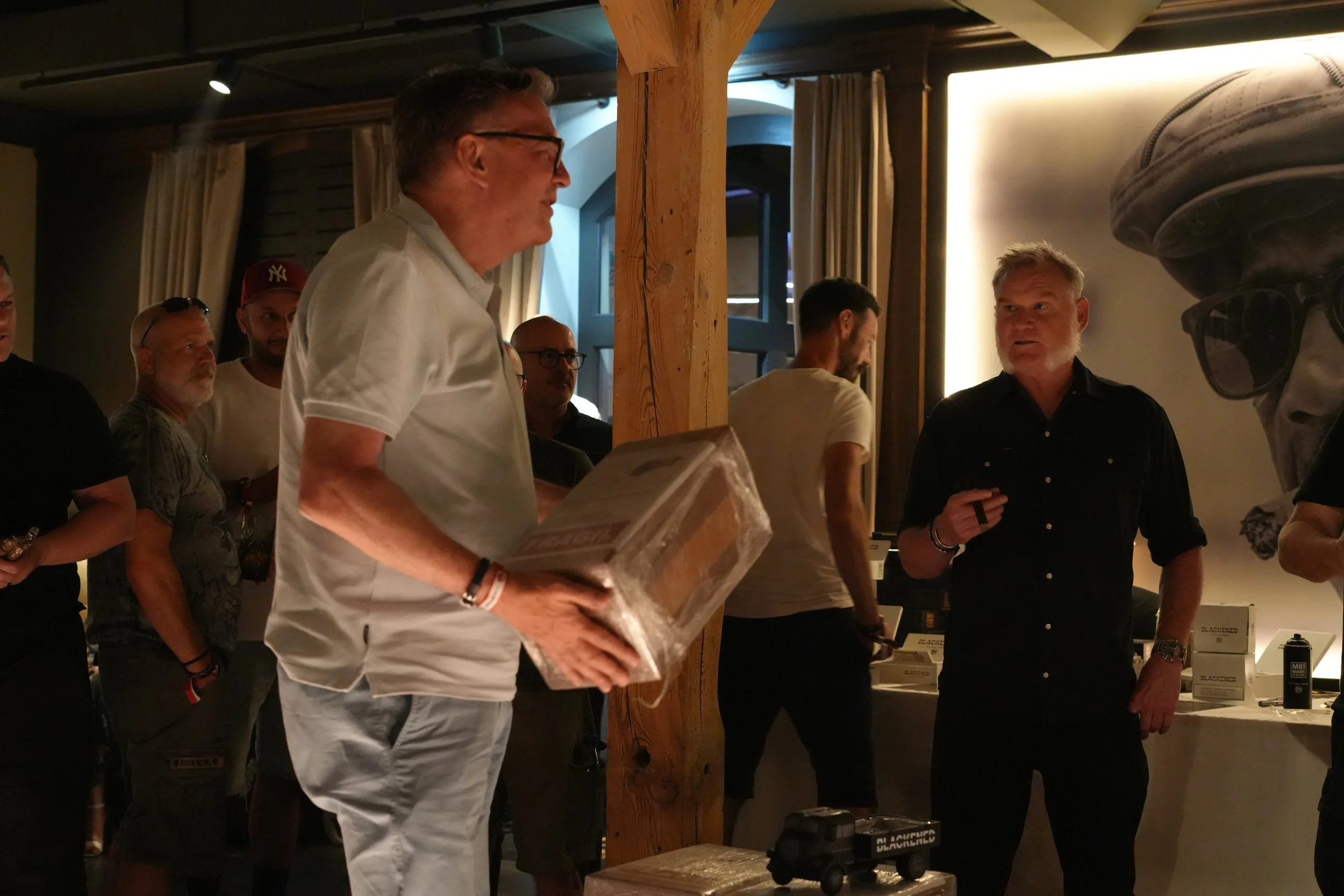 A group of people gathered indoors, with some looking towards a man in a black shirt who appears to be speaking. One man is holding a box, and others are standing near tables with items on display.