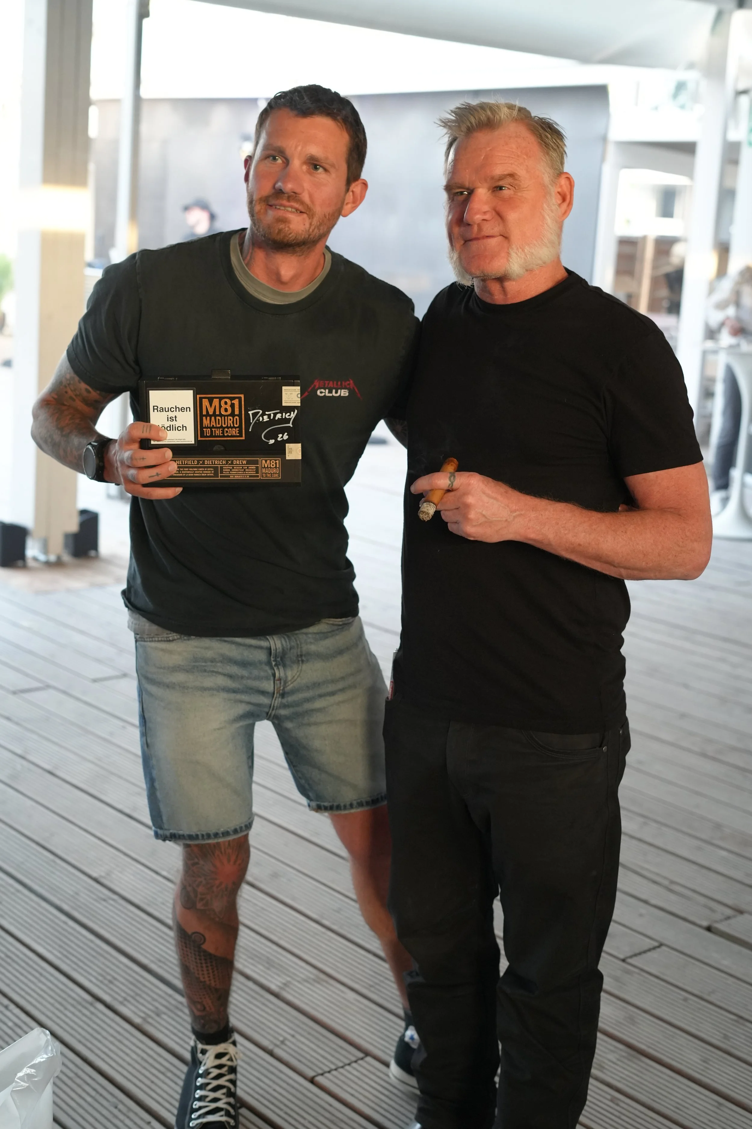 Two men standing on a wooden deck, one man holding a box with warning labels and the other holding a cigar. They are posing for a photo in an outdoor setting with a modern building in the background.