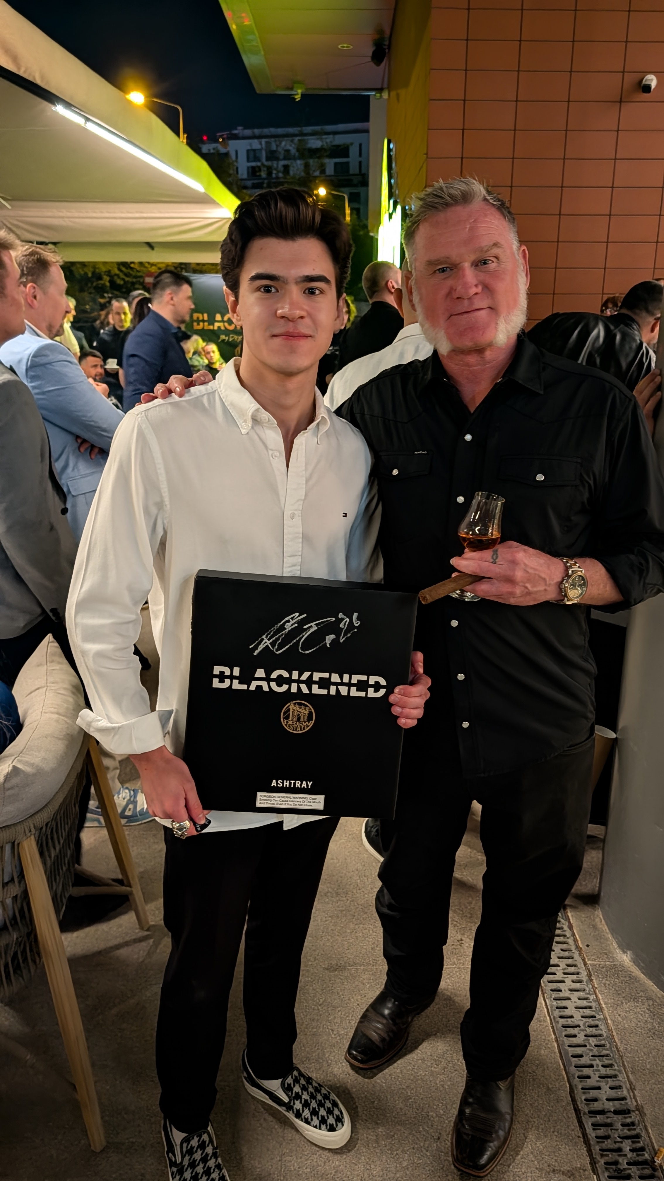 Two men posing together at an event, one holding a framed certificate and the other holding a glass of whiskey. The background shows a crowd of people socializing indoors and outdoors at night.