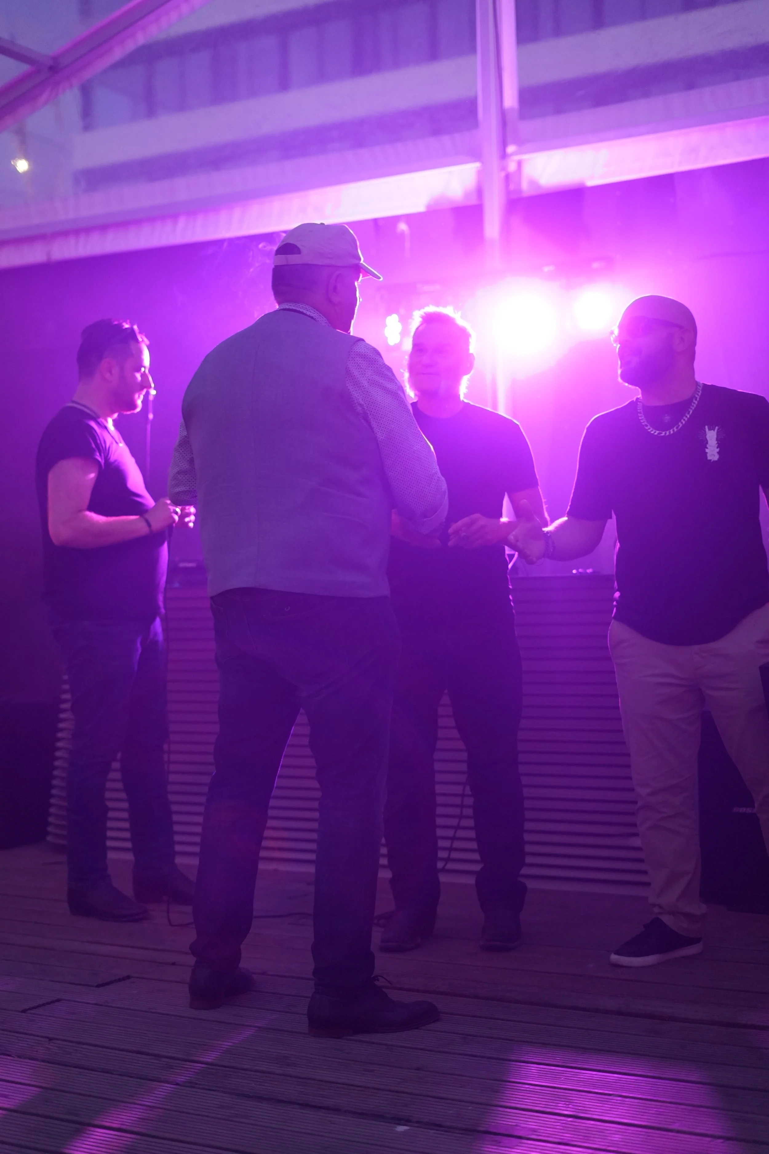 Four men are standing and talking in a dimly lit room with purple lighting. They are casually dressed, with one wearing a hat and a vest, and another with a metallic chain necklace. The floor appears to be wooden.